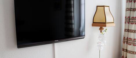 A flat-screen TV mounted on a wall, a table with a remote control, a lamp, and a potted orchid.