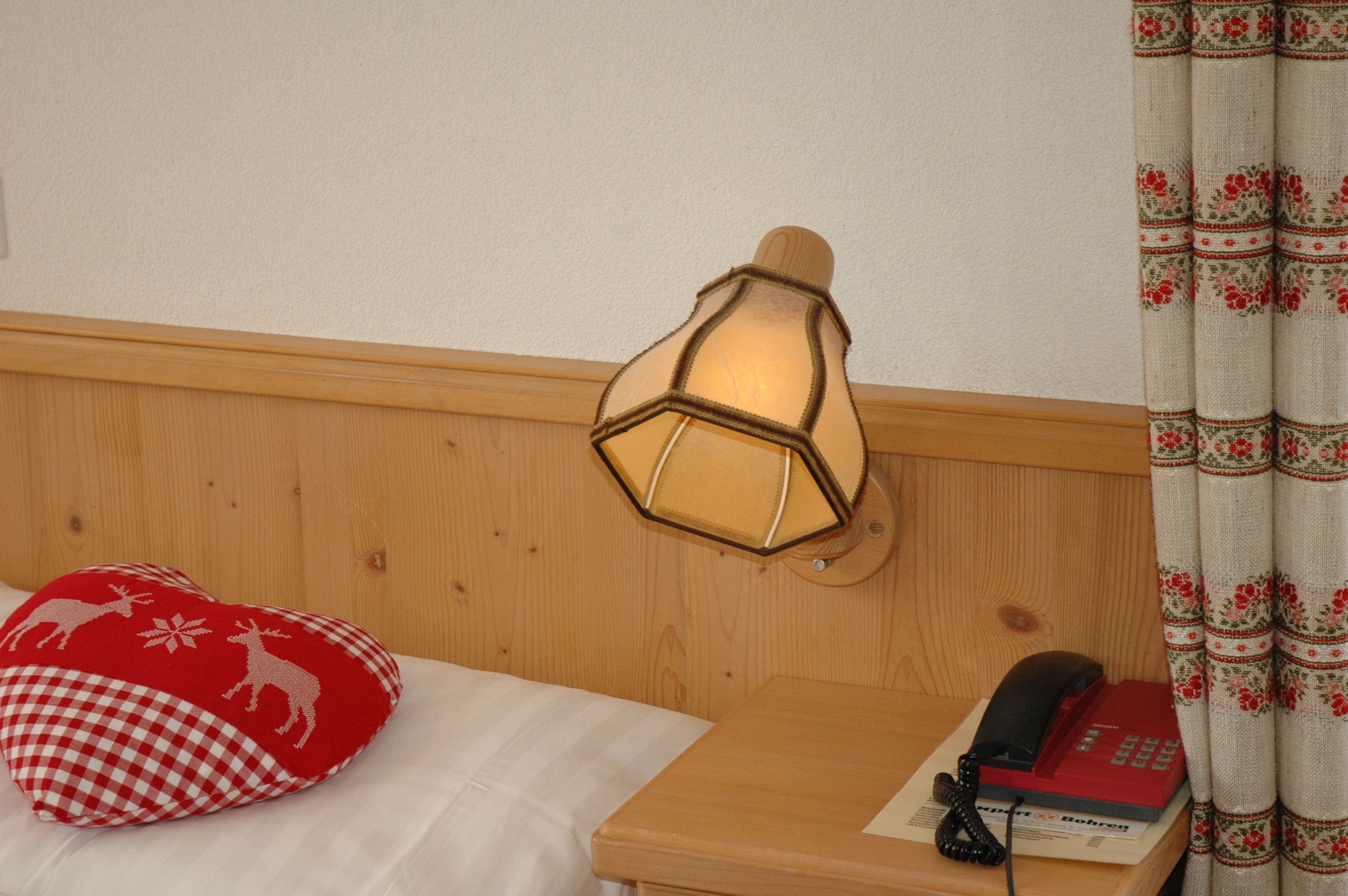 A wooden headboard with a wall-mounted lamp, a red and white checkered pillow, a telephone, and a book on a bedside table.
