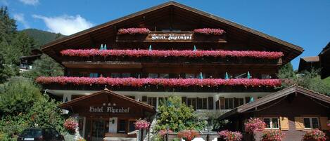 Un hotel de varios pisos con balcones decorados con flores, un letrero que dice "Hotel Alpenhof" y un camino pavimentado al frente.