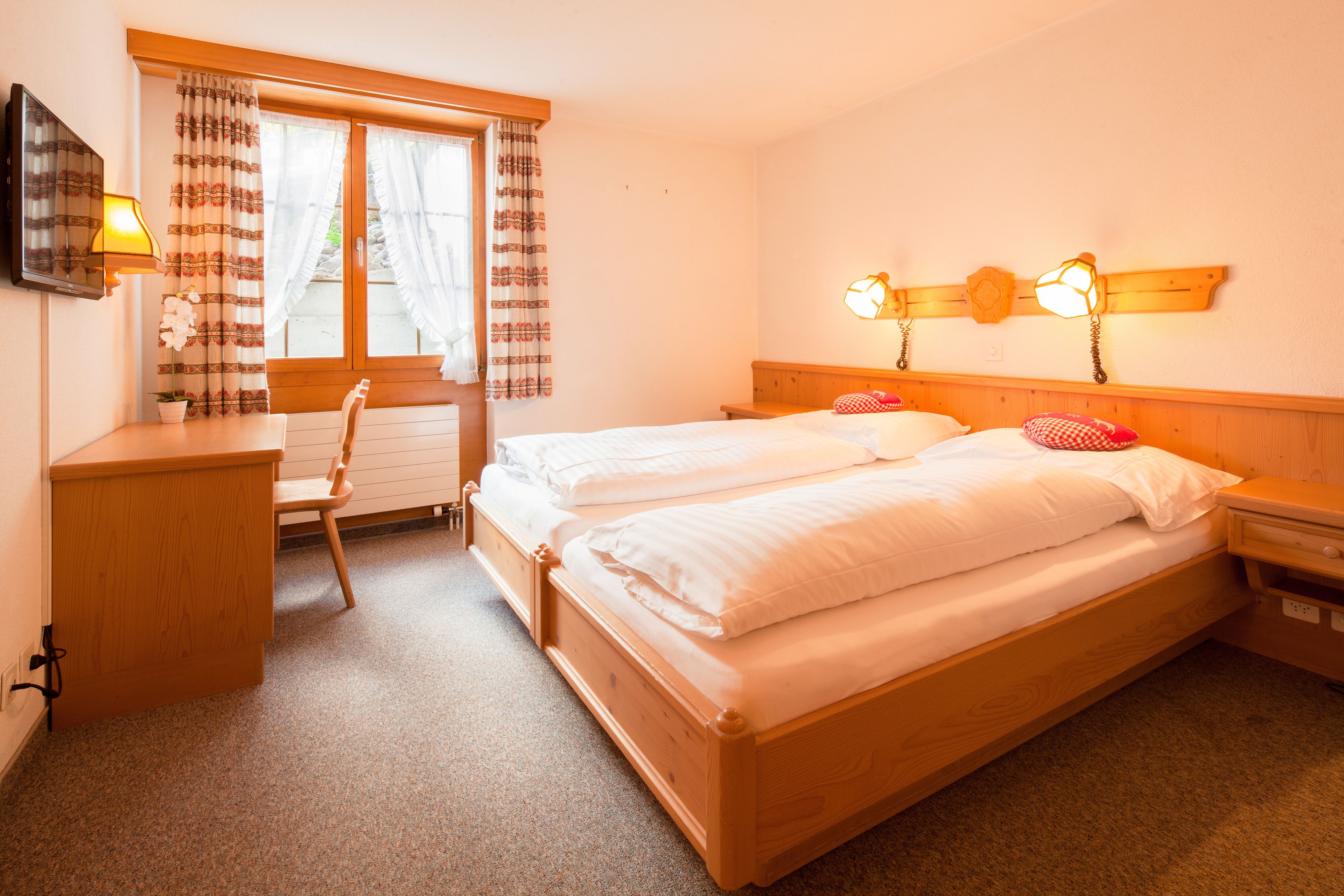 A double bed with wooden headboard, two bedside tables, a desk with a chair, a window with curtains, and a wall-mounted lamp.