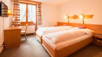 A double bed with wooden headboard, two bedside tables, a desk with a chair, a window with curtains, and a wall-mounted lamp.