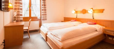 Una cama doble con cabecera de madera, dos mesitas de noche, un escritorio con silla, una ventana con cortinas y una lámpara de pared.