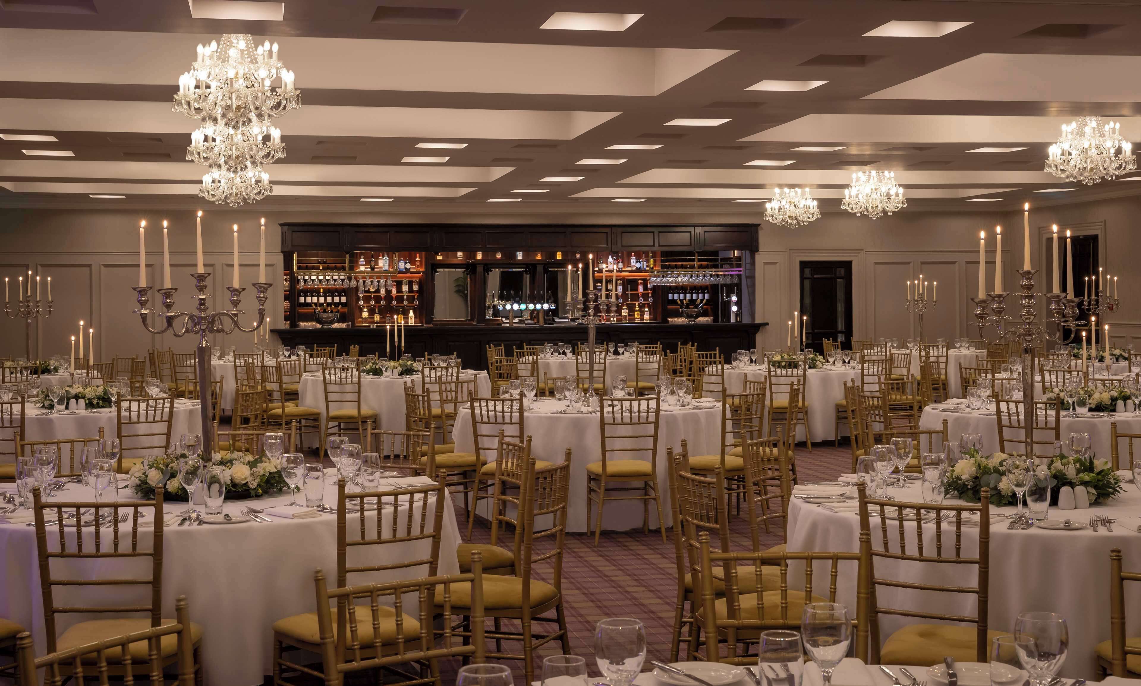 Een feestzaal met ronde tafels, gouden stoelen en kroonluchters.