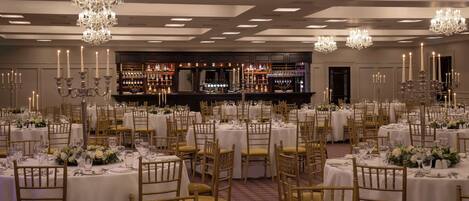 A banquet hall with round tables, gold chairs, and chandeliers.