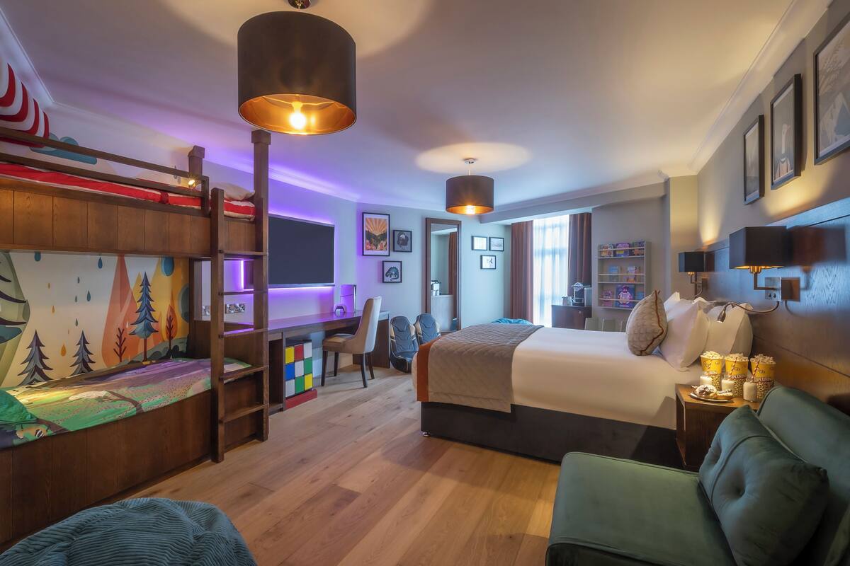 A hotel room with a bunk bed, a desk, a chair, a bed, a couch, and a wall with framed pictures.