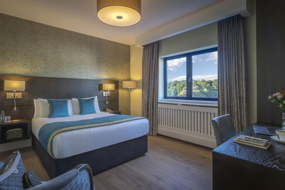 A hotel room with a large bed, bedside lamps, a desk with a laptop and coffee, and a window with a view of trees.