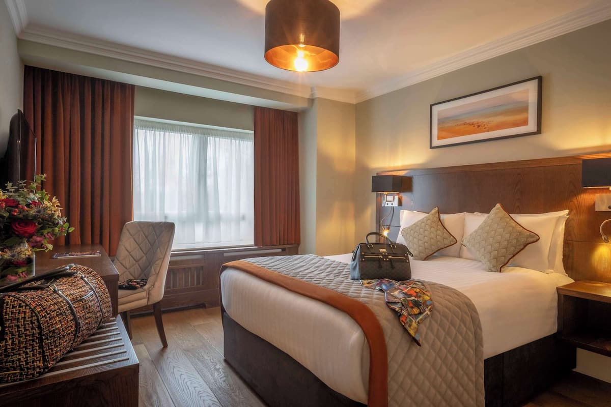 A hotel room with a large bed, a desk with a chair, a window with curtains, a painting on the wall, and a lamp on the nightstand.