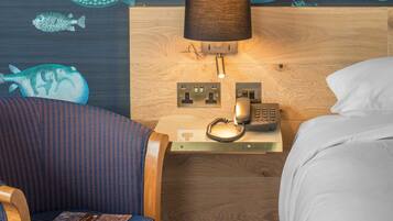 A hotel room with a wooden headboard, a bedside table with a lamp, a telephone, and a wall with fish patterns.