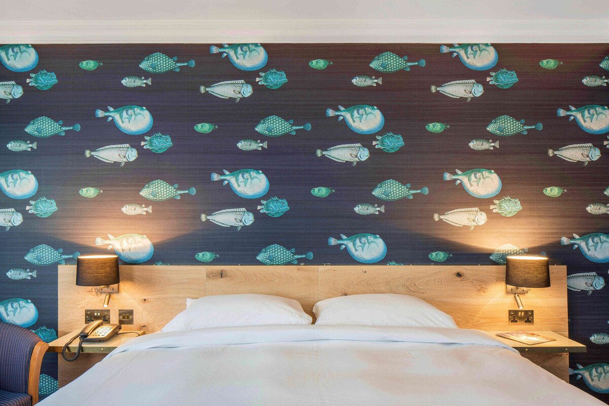 A hotel room with a bed, two bedside lamps, a telephone, and a wall with a fish pattern.