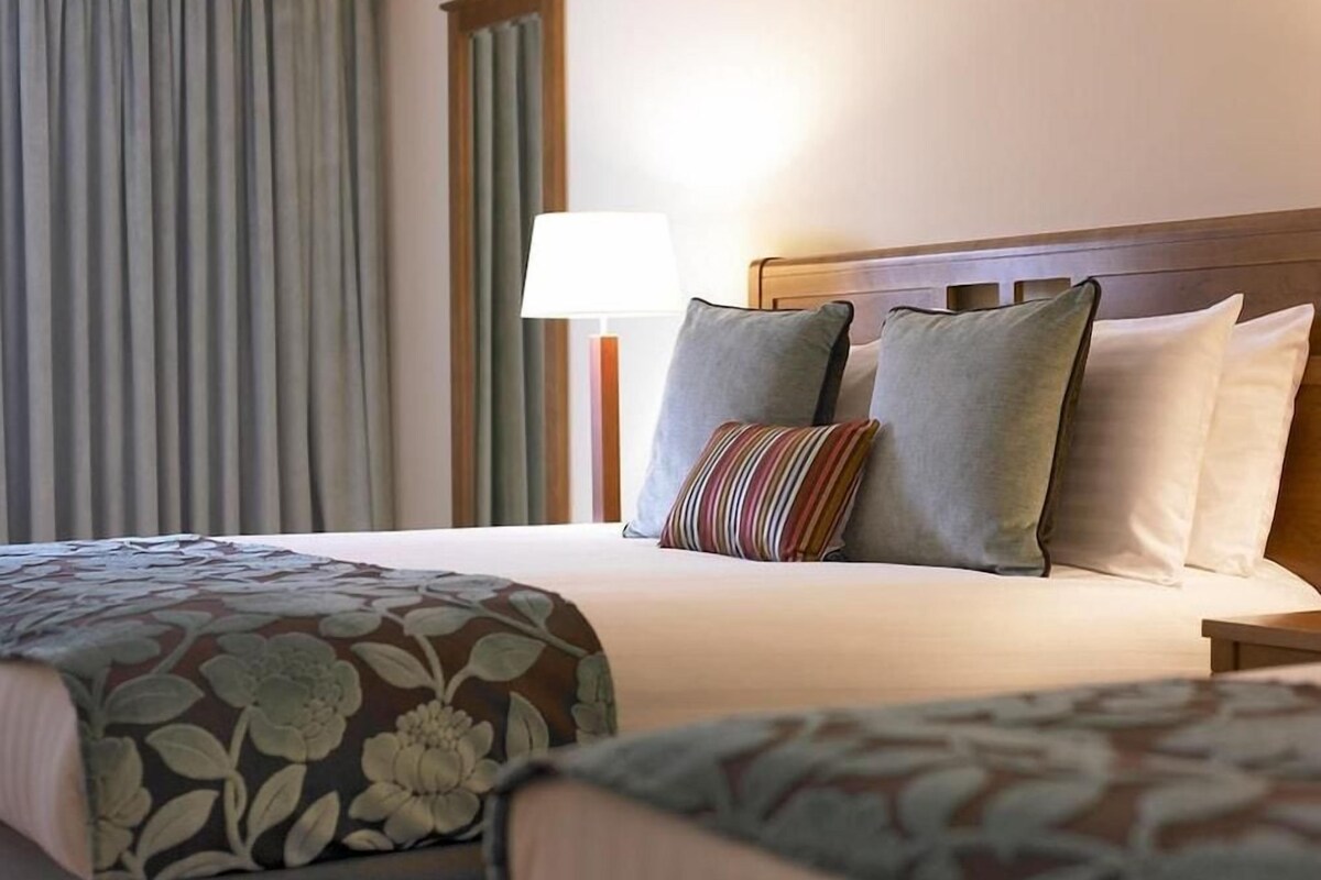 Una habitación de hotel con una cama, una mesita de noche, una lámpara y una ventana con cortinas.