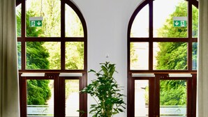 Un vestíbulo con dos grandes ventanales de arco, una jardinera de madera con una planta, un detector de incendios y una recepción con una silla.