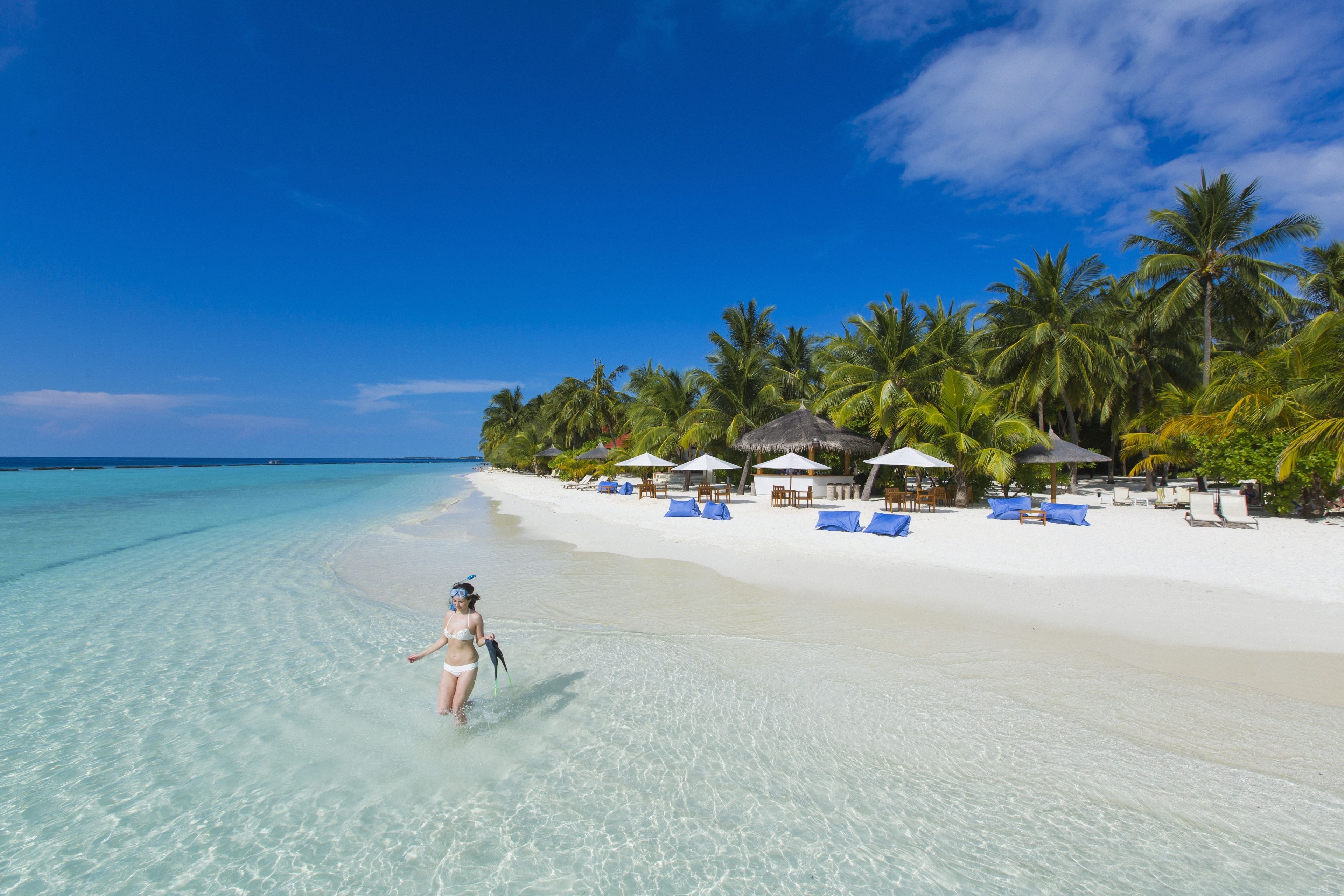 A beach with clear turquoise water, a woman in a bikini, and a resort with palm trees and lounge chairs.