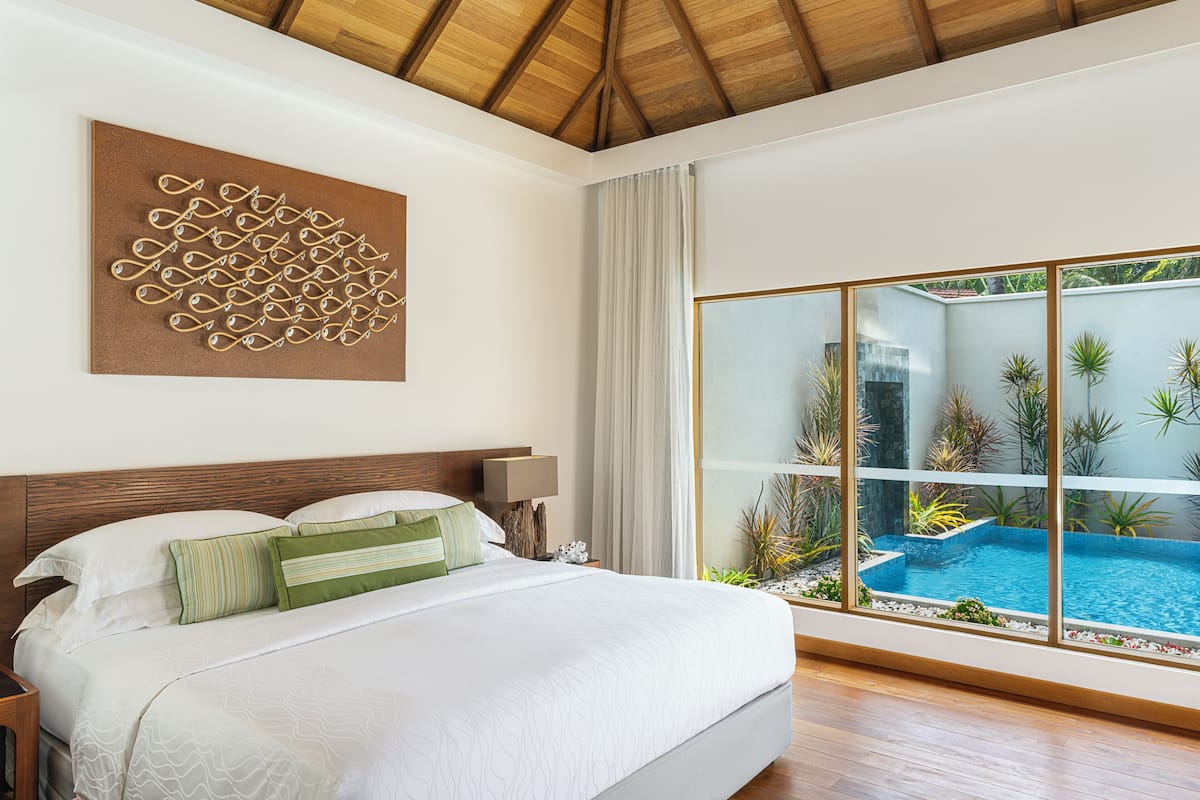 Camera da letto con un letto grande, soffitto in legno, vista su una piscina e un elemento decorativo a parete.