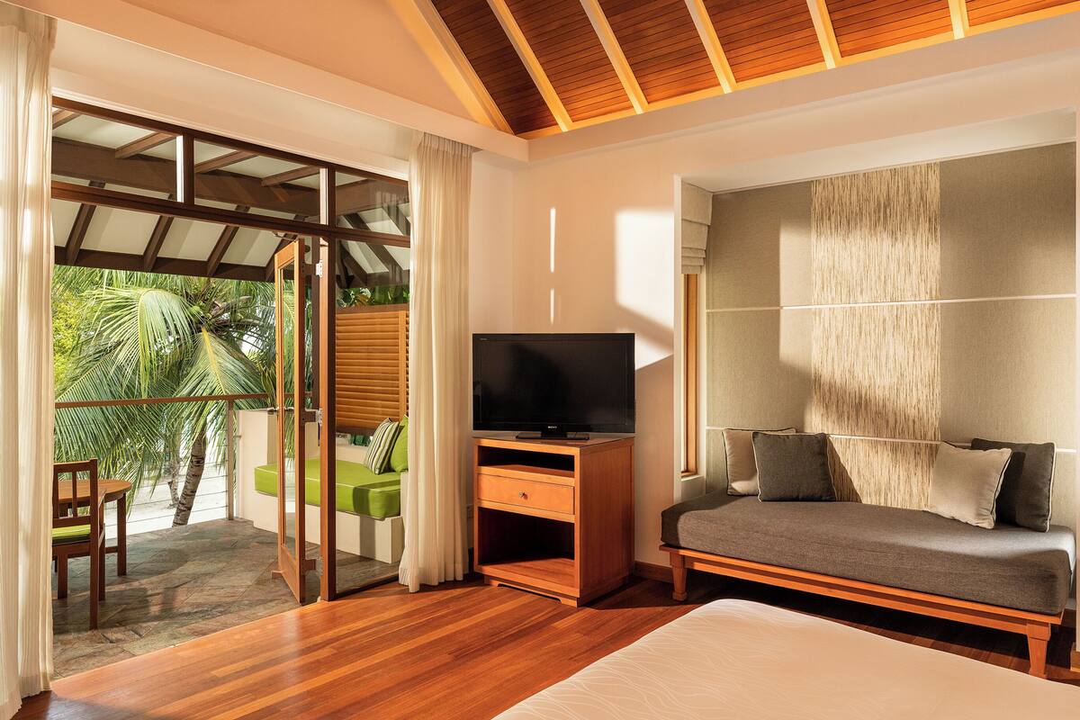 A hotel room with a wooden floor, a sofa, a TV on a wooden stand, and a balcony with a view of palm trees.