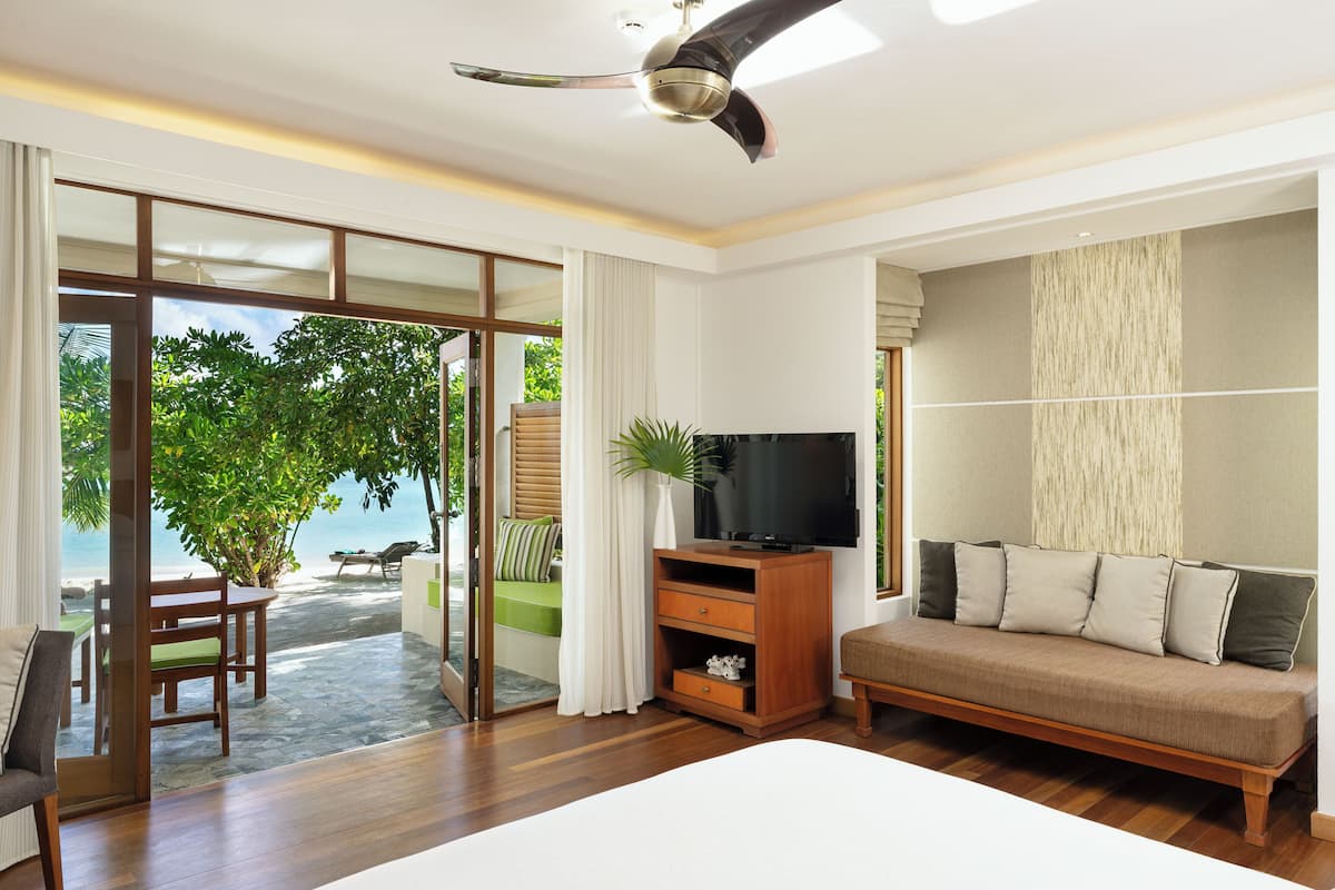 A spacious room with a large bed, a TV on a wooden stand, and a sliding glass door leading to a balcony with a view of the ocean.