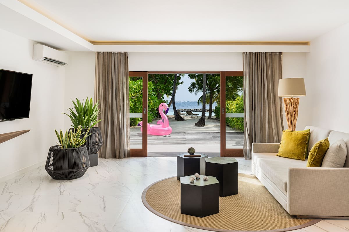 A modern living room with a sofa, a TV, and a view of a beach with a flamingo float.