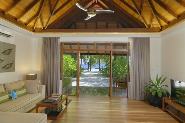 A spacious living area with a wooden ceiling, a large TV, a sofa, and a view of a beach.