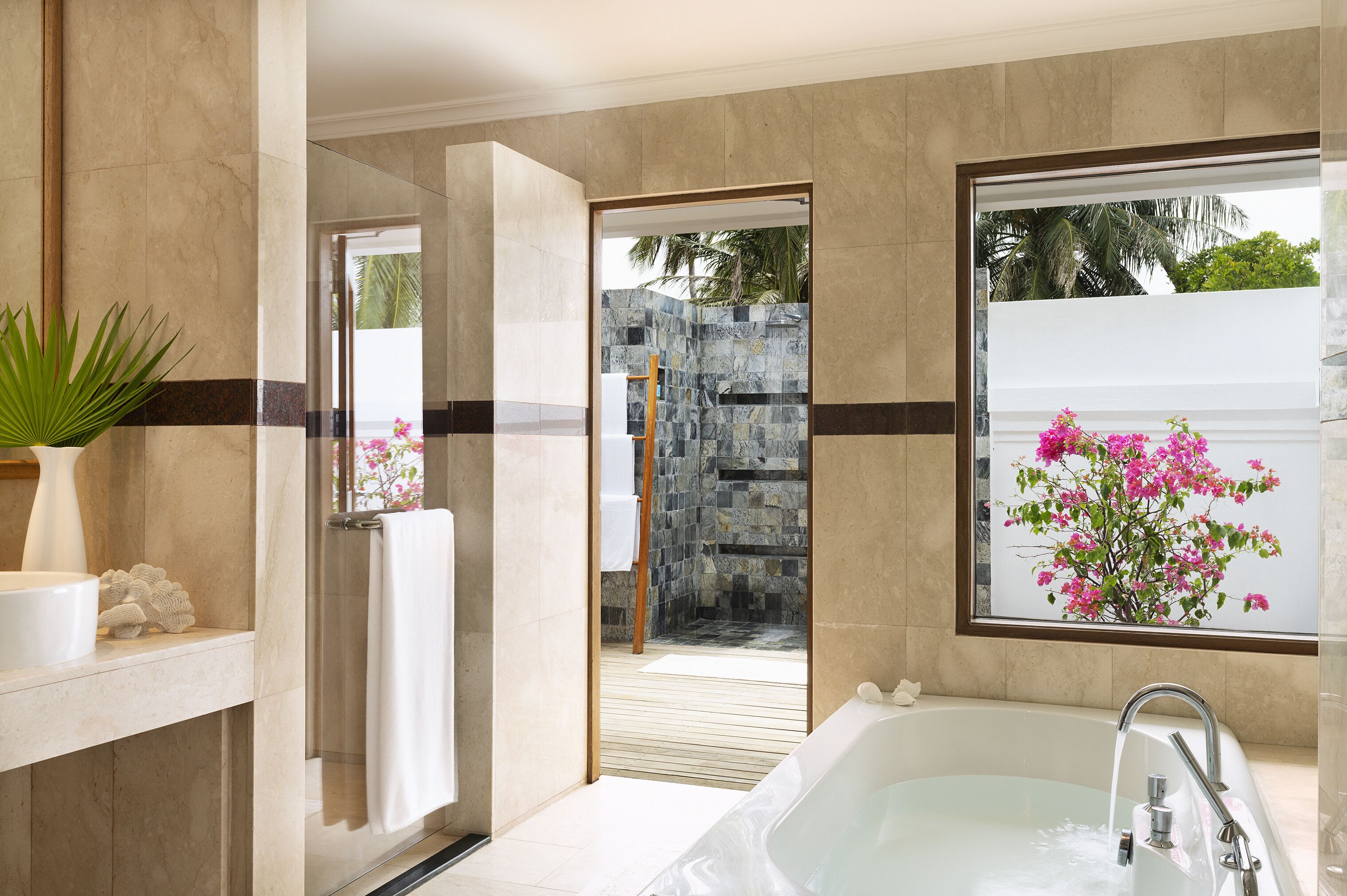 A modern bathroom with a bathtub, a shower, and a large window with a view of a stone wall and tropical plants.