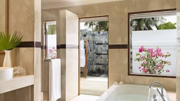 A modern bathroom with a bathtub, a shower, and a large window with a view of a stone wall and tropical plants.