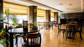 Um restaurante espaçoso com pé-direito alto, mesas e cadeiras de madeira e amplas janelas com persianas.