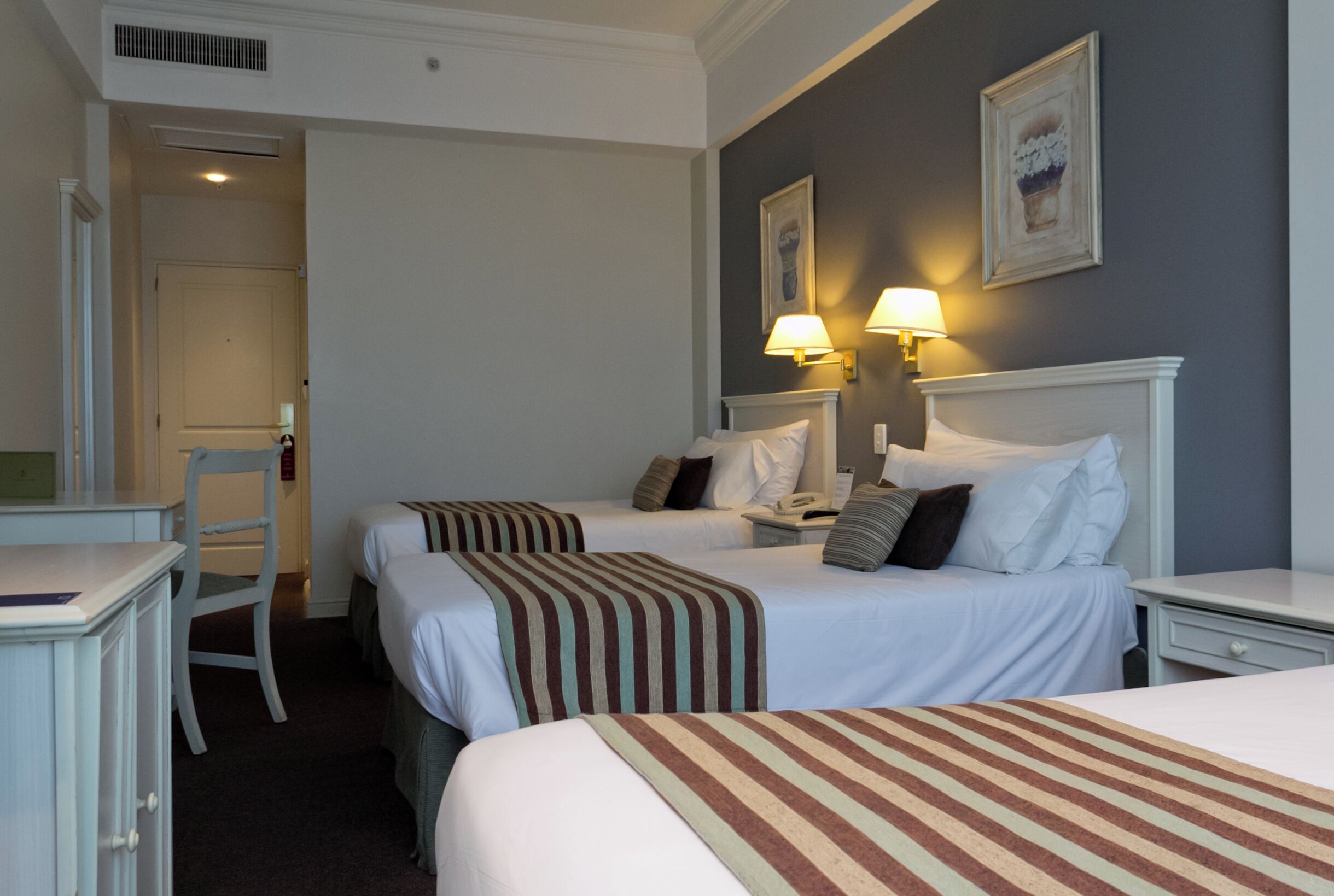 A hotel room with two beds, a desk, and a chair. There are two bedside lamps and a framed picture on the wall.