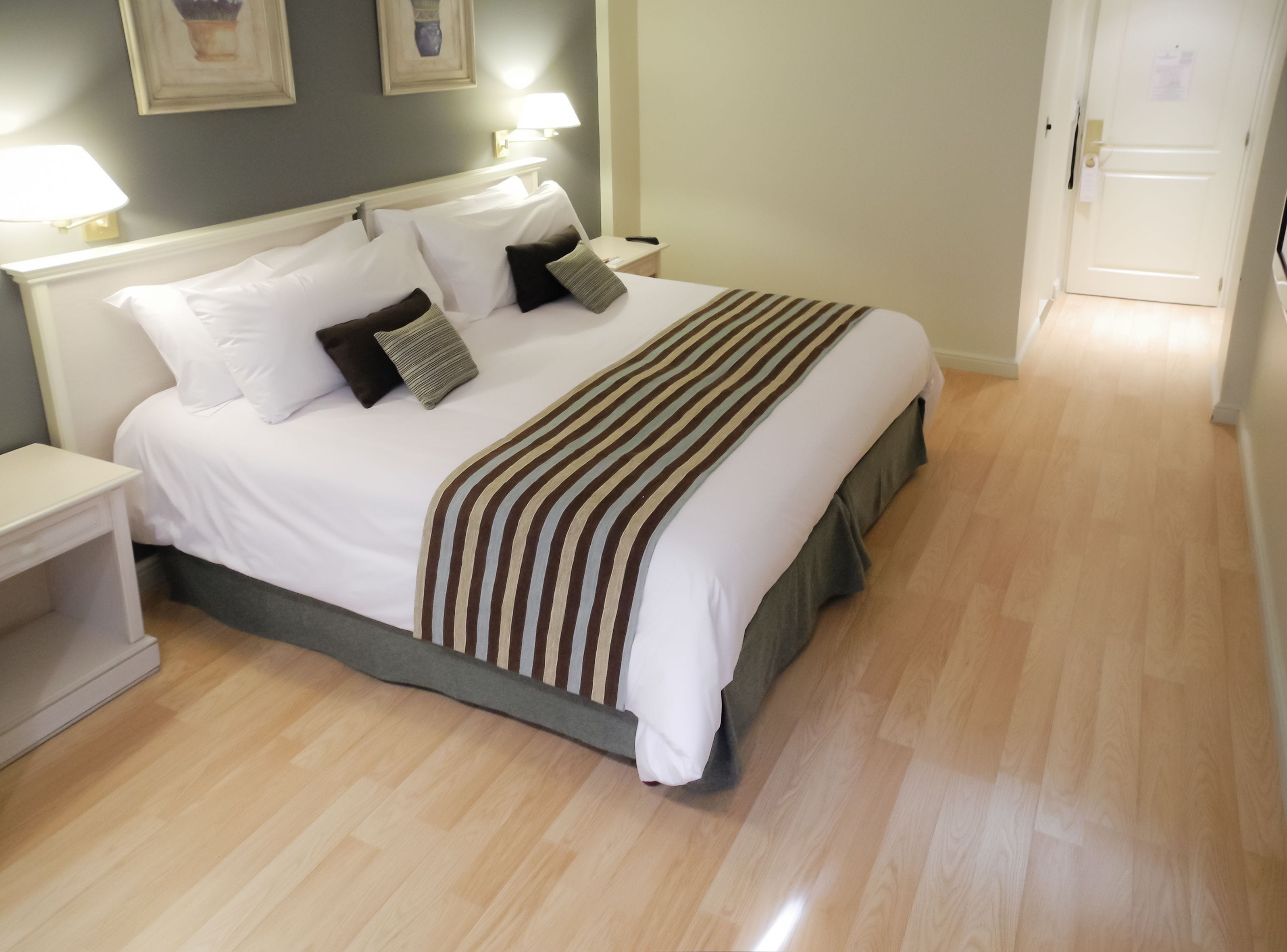A neatly made bed with a striped blanket, white pillows, and a grey skirt. There is a nightstand with a lamp and a wall-mounted light. The room has wooden flooring and a door in the background.