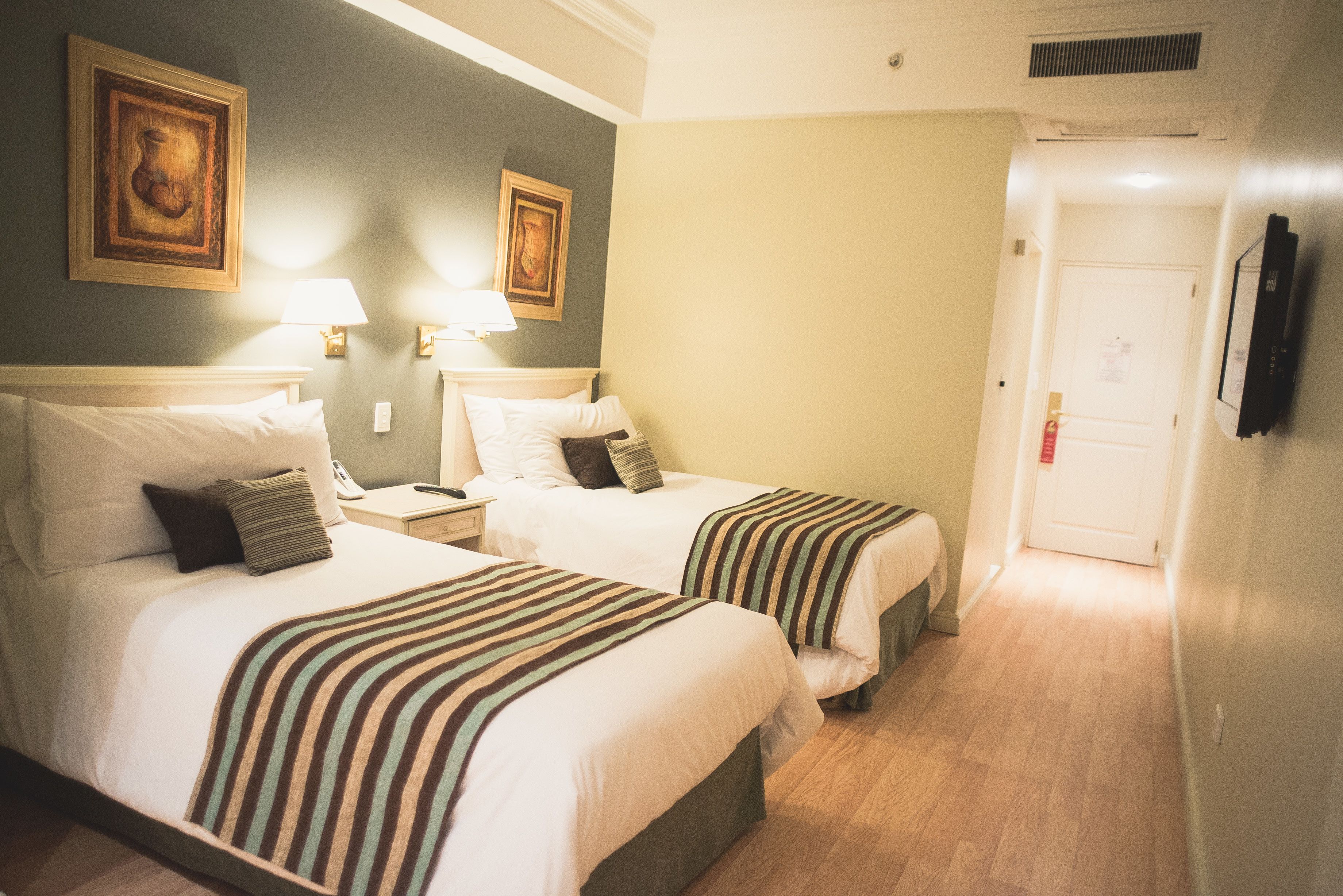 A hotel room with two beds, a nightstand with a lamp, and a door leading to another room.