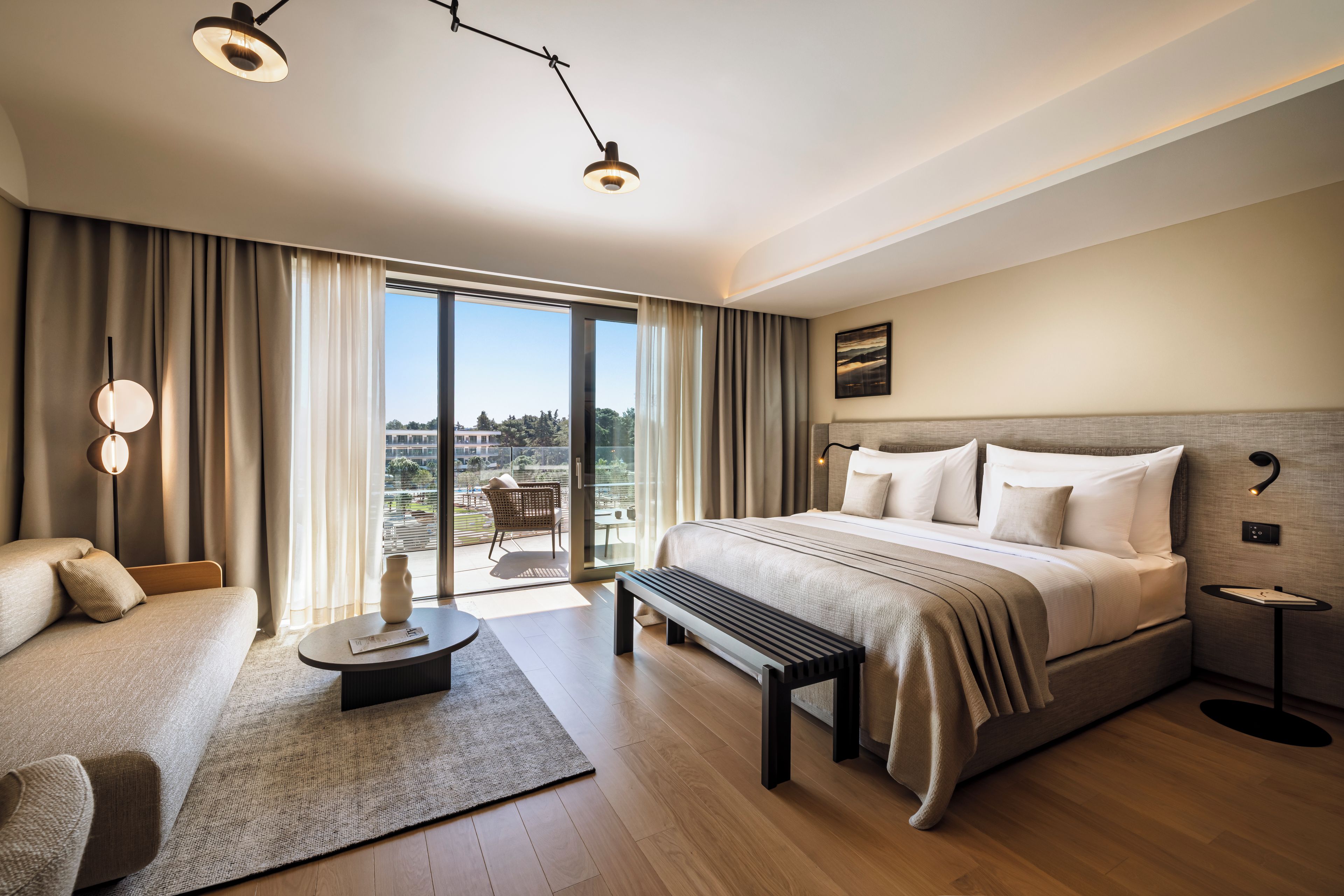 Una habitación de hotel moderna con una cama grande, un sofá, un banco y vistas de la ciudad a través del balcón.