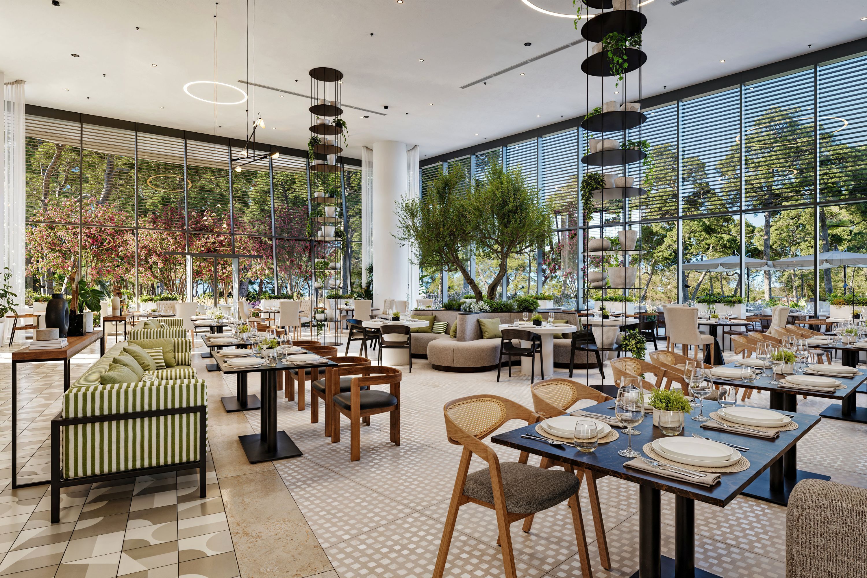 A spacious indoor dining area with multiple tables and chairs, large windows offering views of trees and sky, and hanging plants.