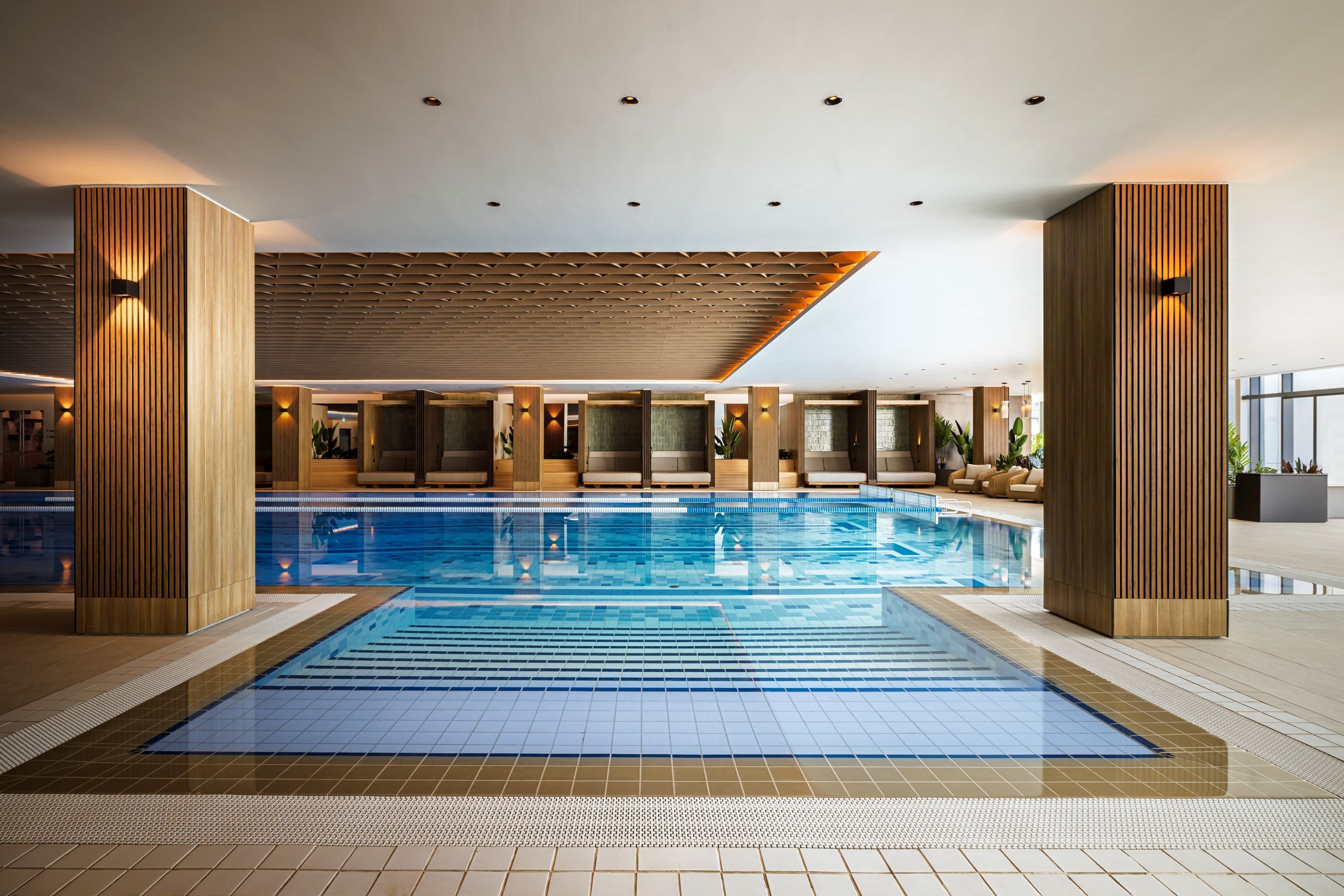 A swimming pool with a tiled floor, wooden paneling, and seating areas with plants.