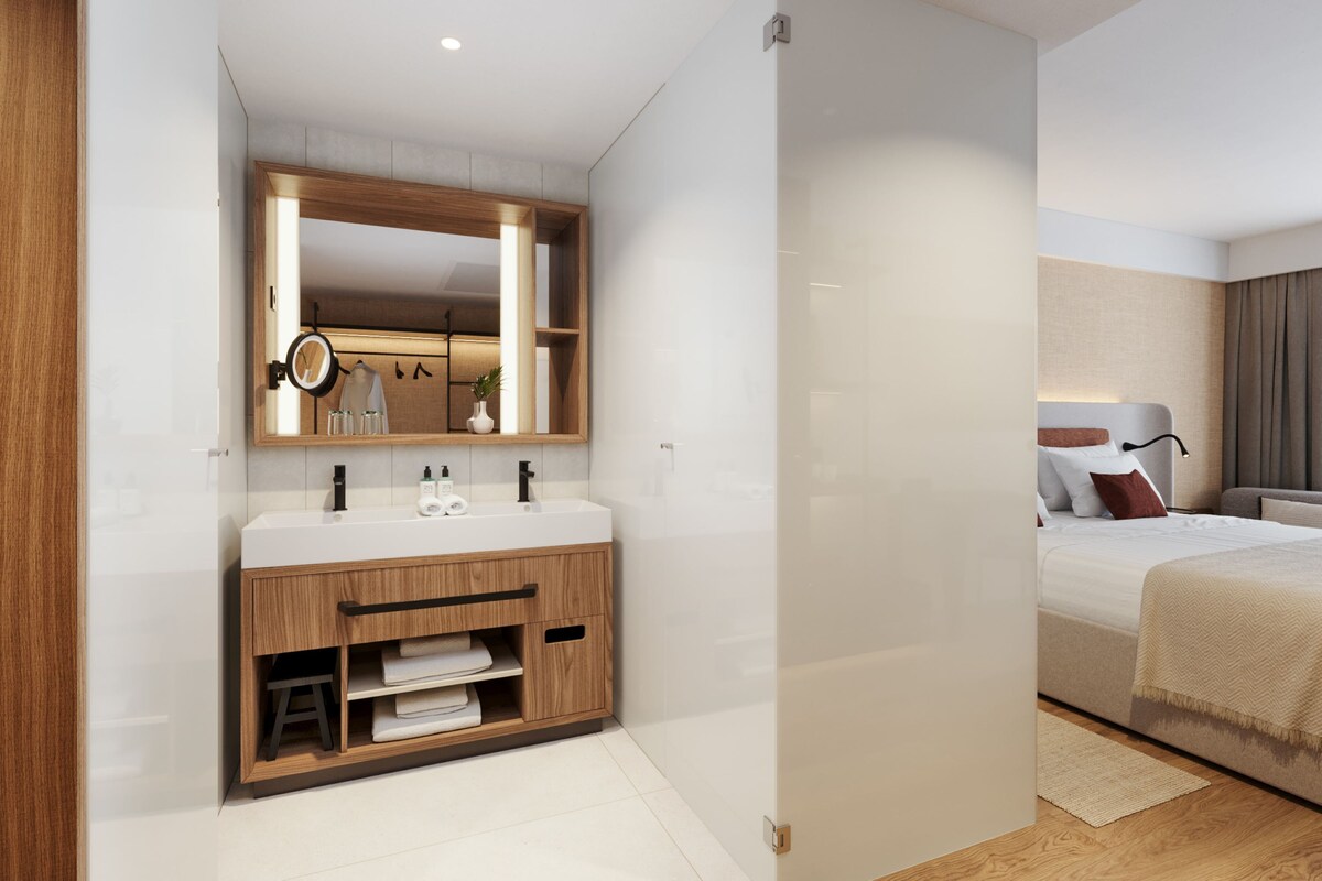 A modern bathroom with a wooden vanity, a large mirror, and a bed in the adjacent room.