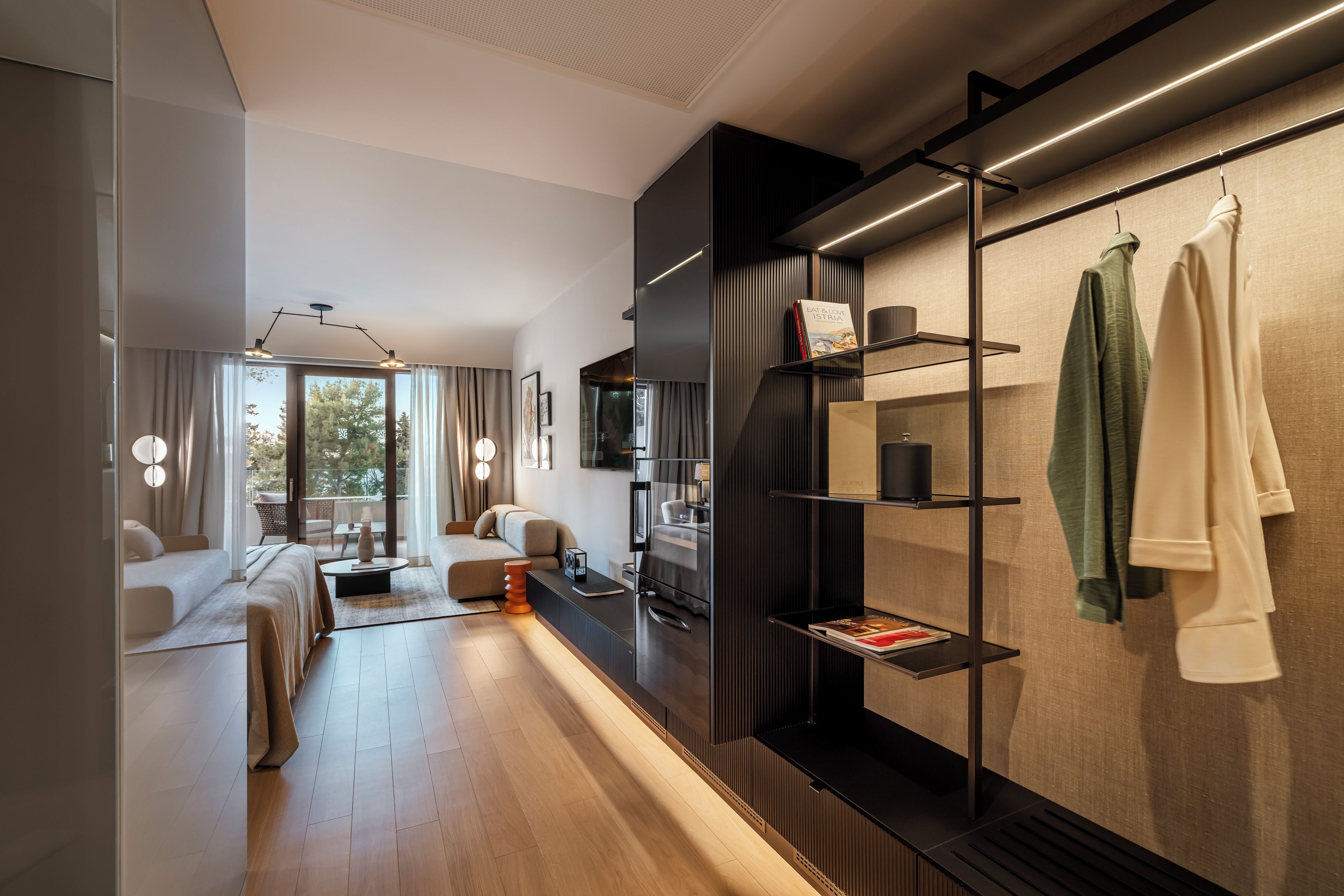 A modern hotel room with a sofa, a dining table, a bookshelf, and a view of the outdoors.