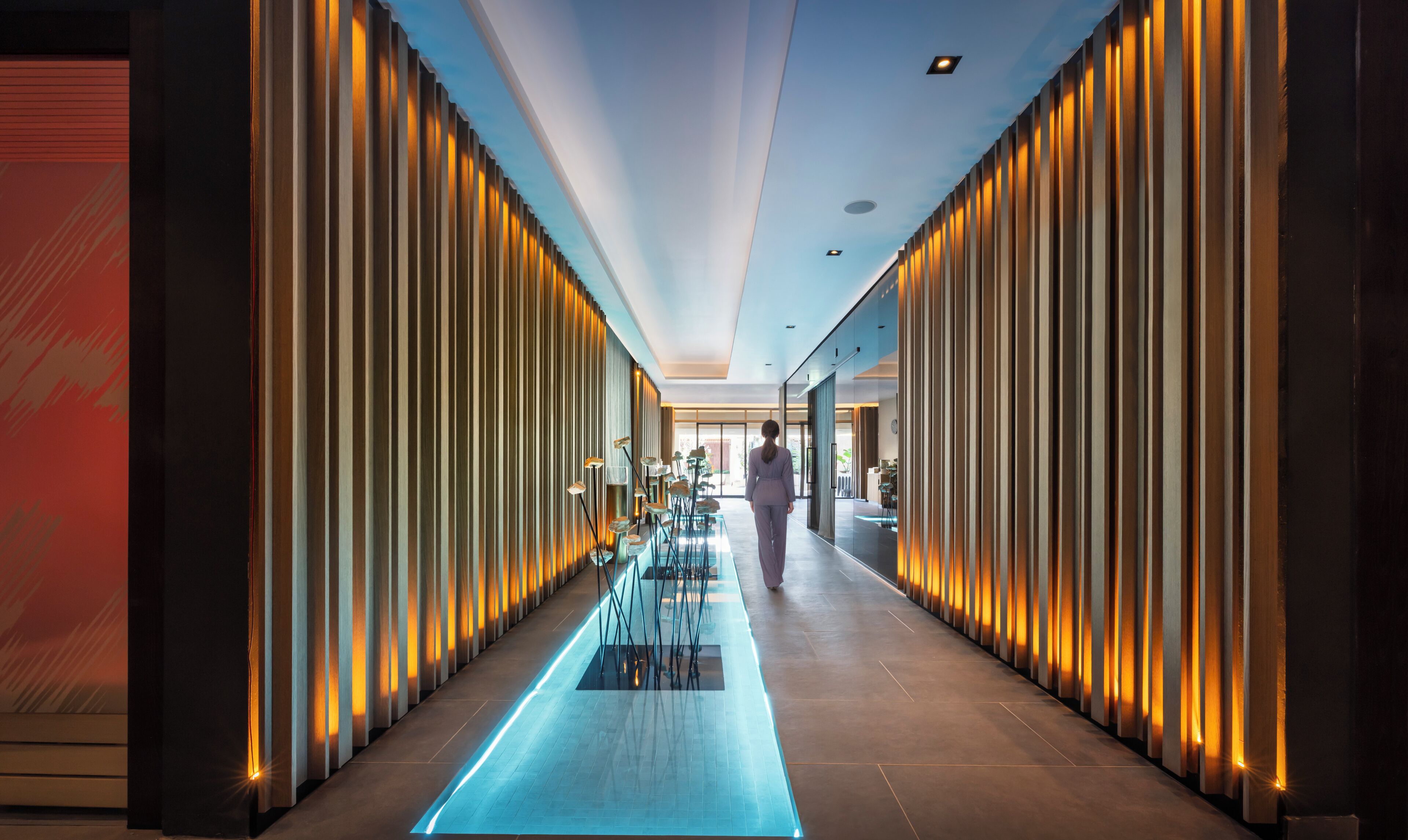 A modern hotel corridor with a central walkway, flanked by vertical light panels and a glass partition.