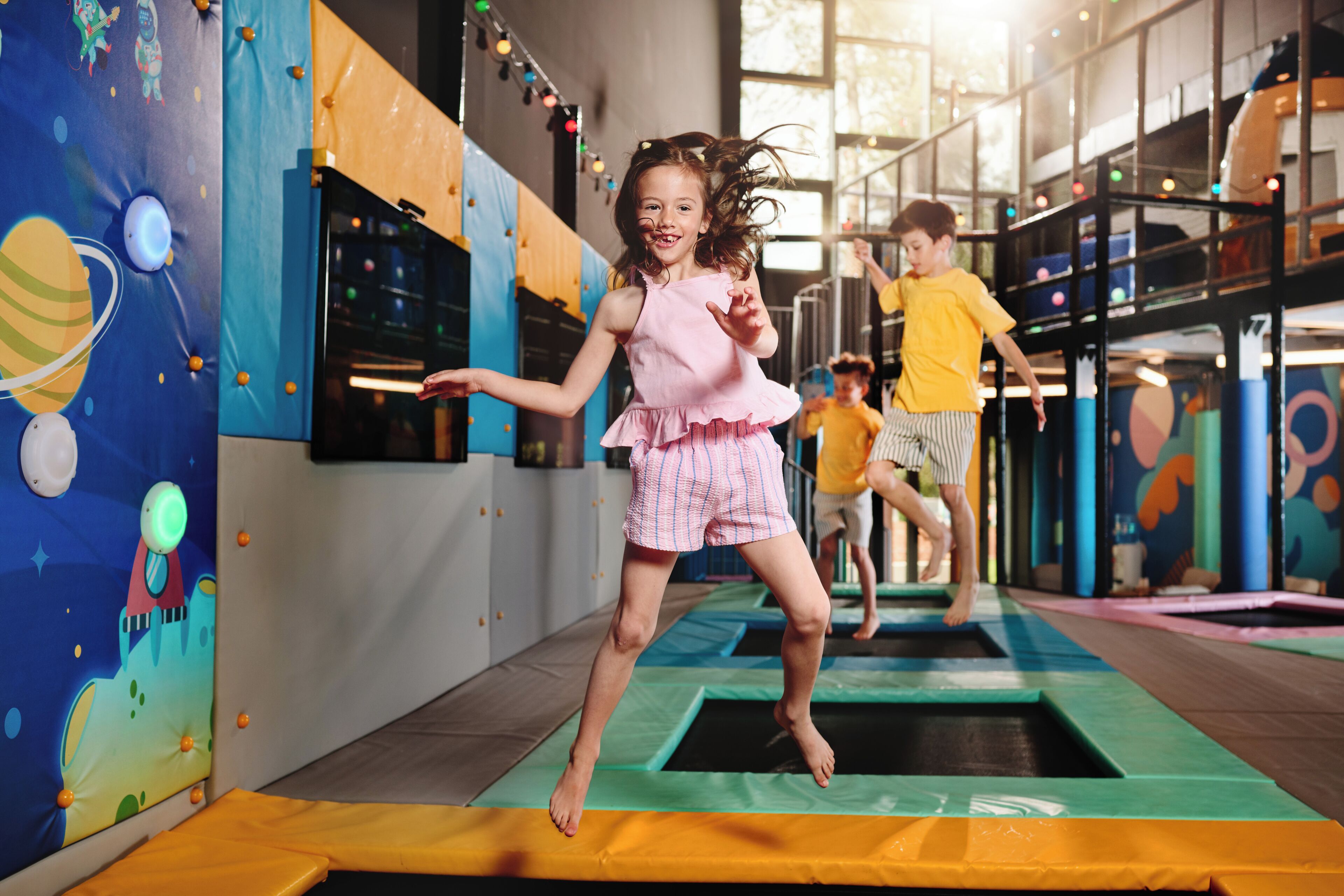 A child jumping on a trampoline indoors with colorful wall decorations and other children in the background.