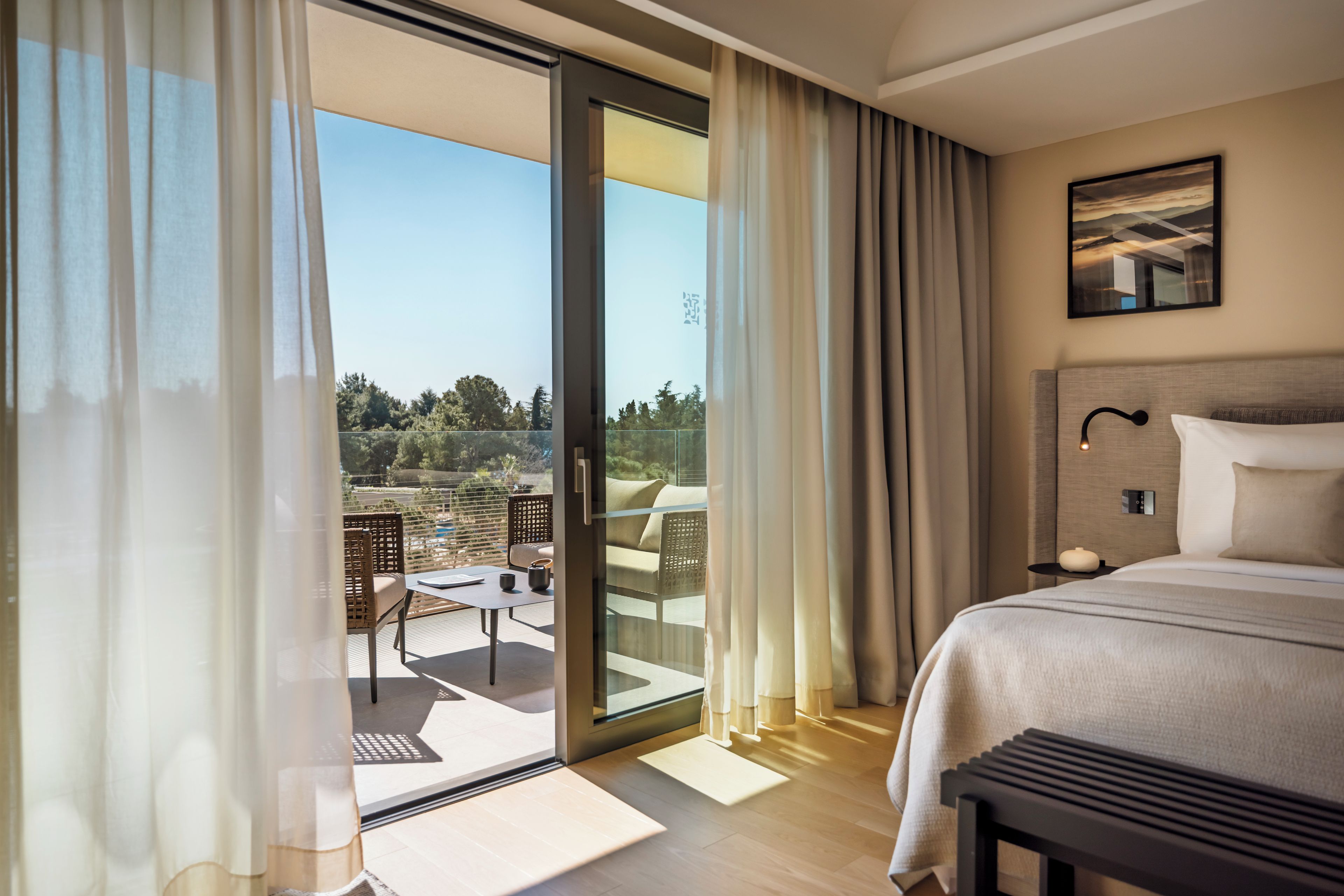 Een hotelkamer met een bed, een nachtkastje, een schilderij aan de muur en uitzicht op de omgeving via een schuifdeur.