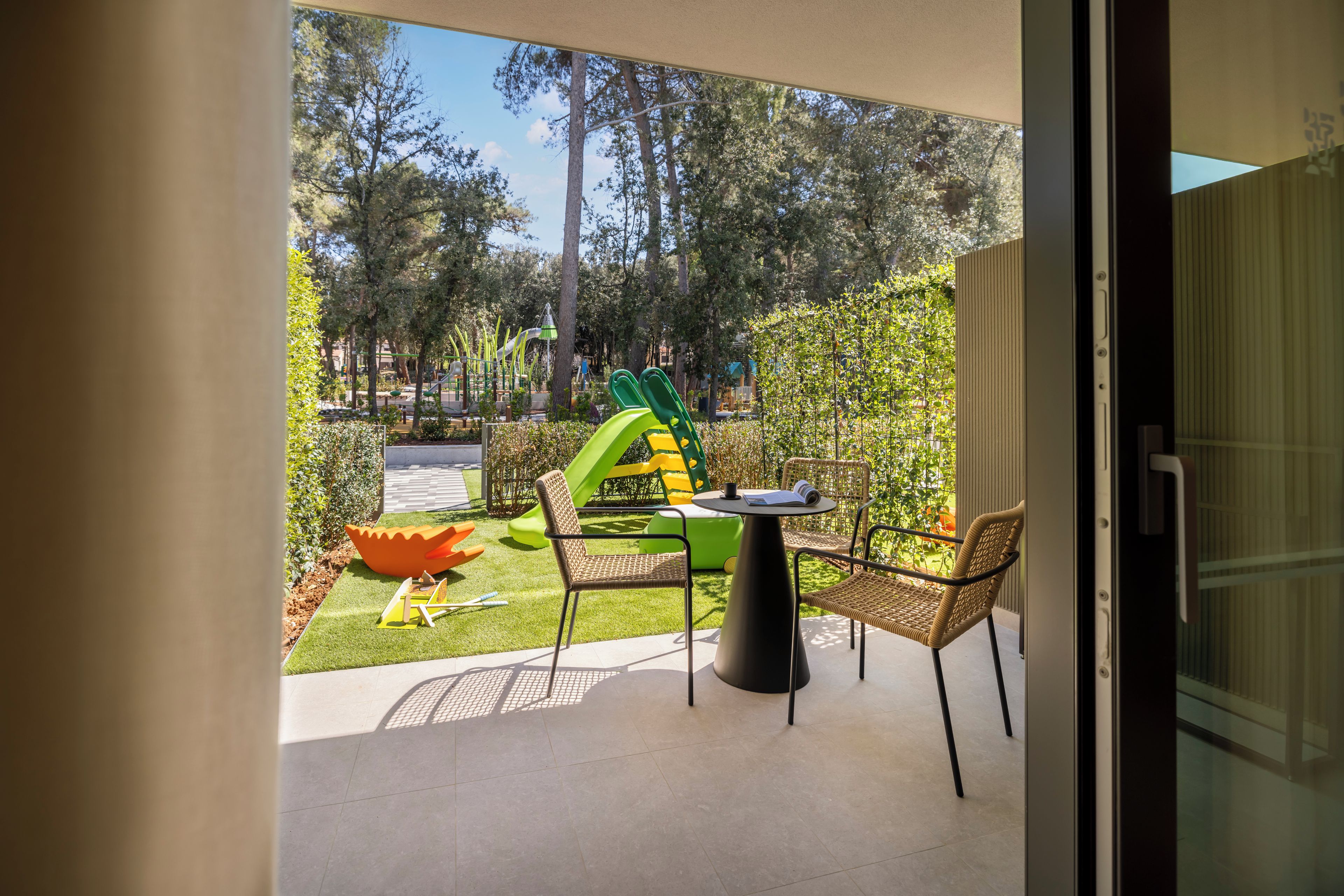Um pátio com mesa e cadeiras, área de recreação infantil com escorregadores e estruturas de escalada, e vista para um parque com árvores e balneário.