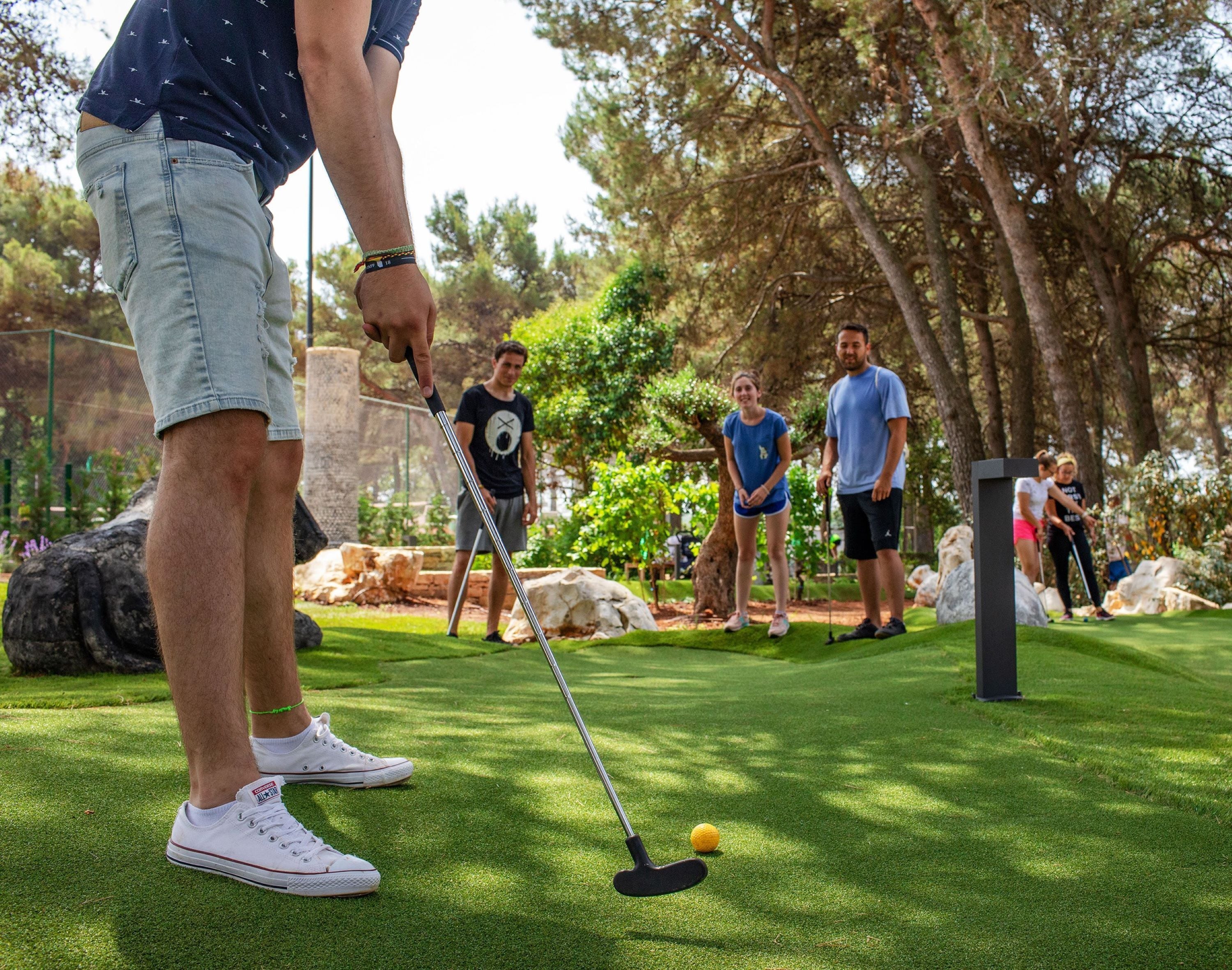 A person preparing to put a golf ball on a putting green, with others standing nearby in a park-like setting.