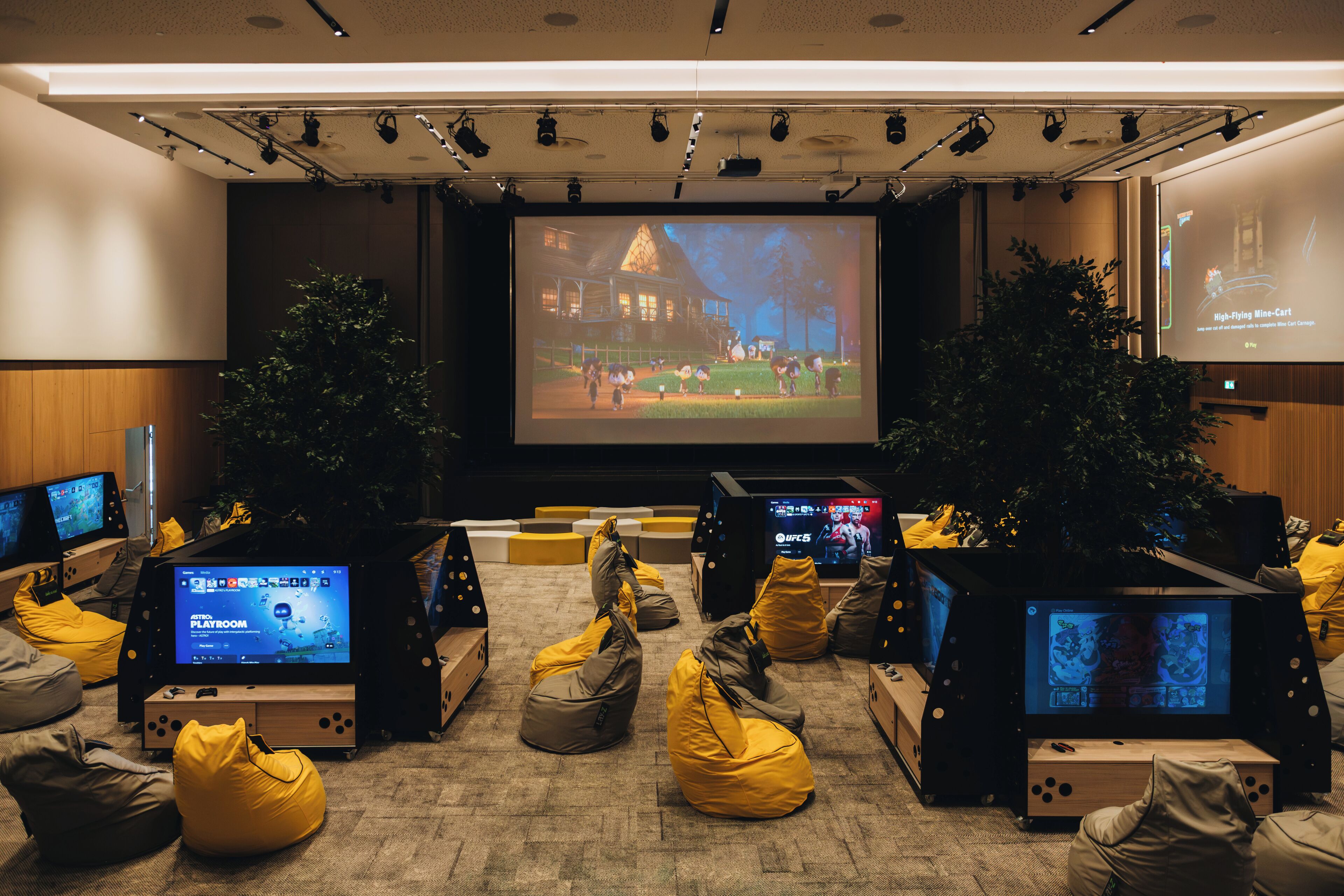 A modern gaming lounge with multiple screens, bean bag seating, and a large projection screen displaying a game.