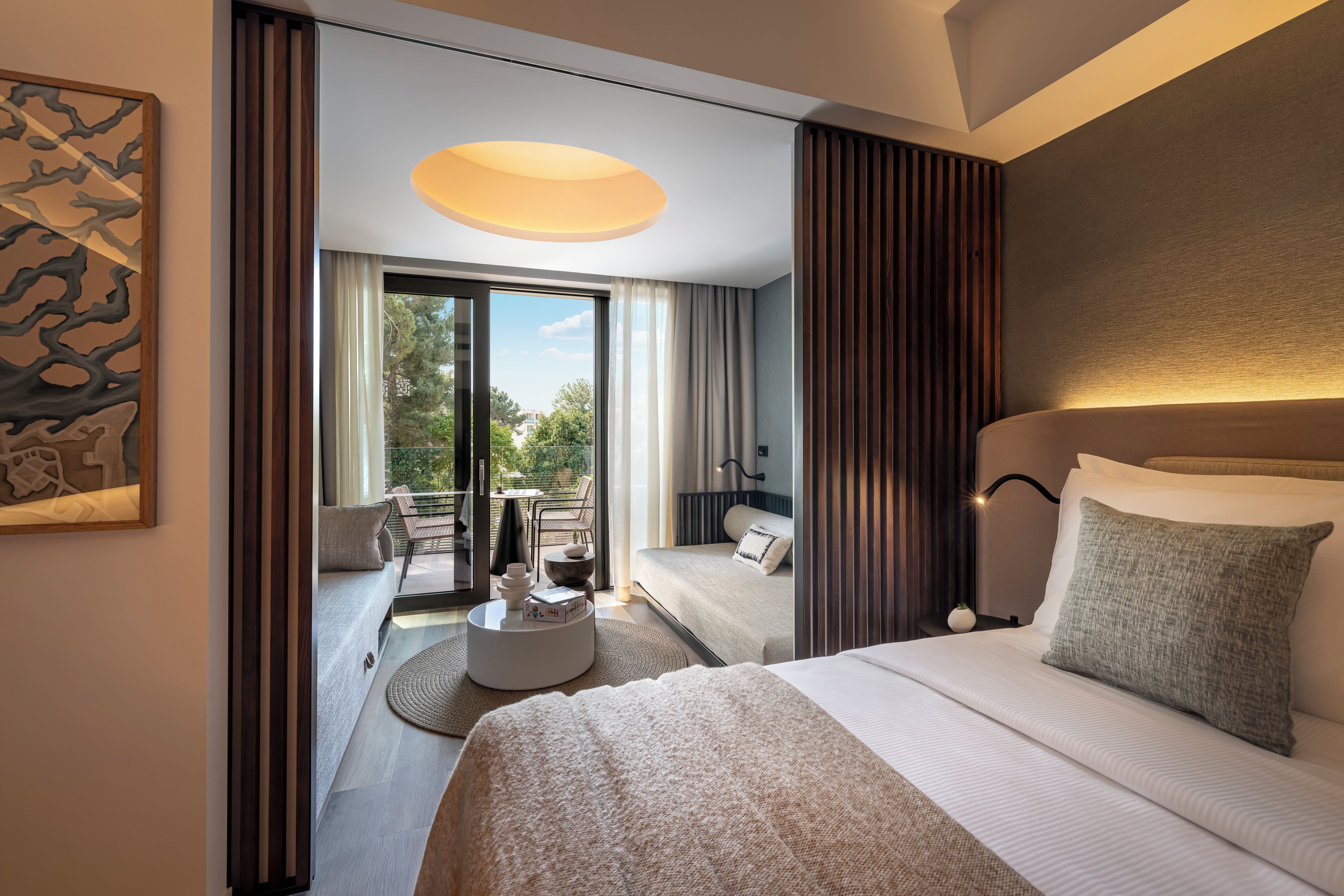 A modern hotel room with a large bed, a balcony with outdoor furniture, and a view of the outdoors.