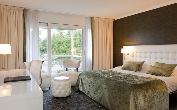 A hotel room with a large bed, a chair, a small ottoman, and a desk with a lamp. - Van der Valk De Gouden Leeuw (Voorschoten)