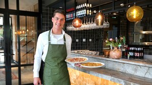 A person in a green apron holding plates of food at a bar with a tiled backsplash and hanging light fixtures.