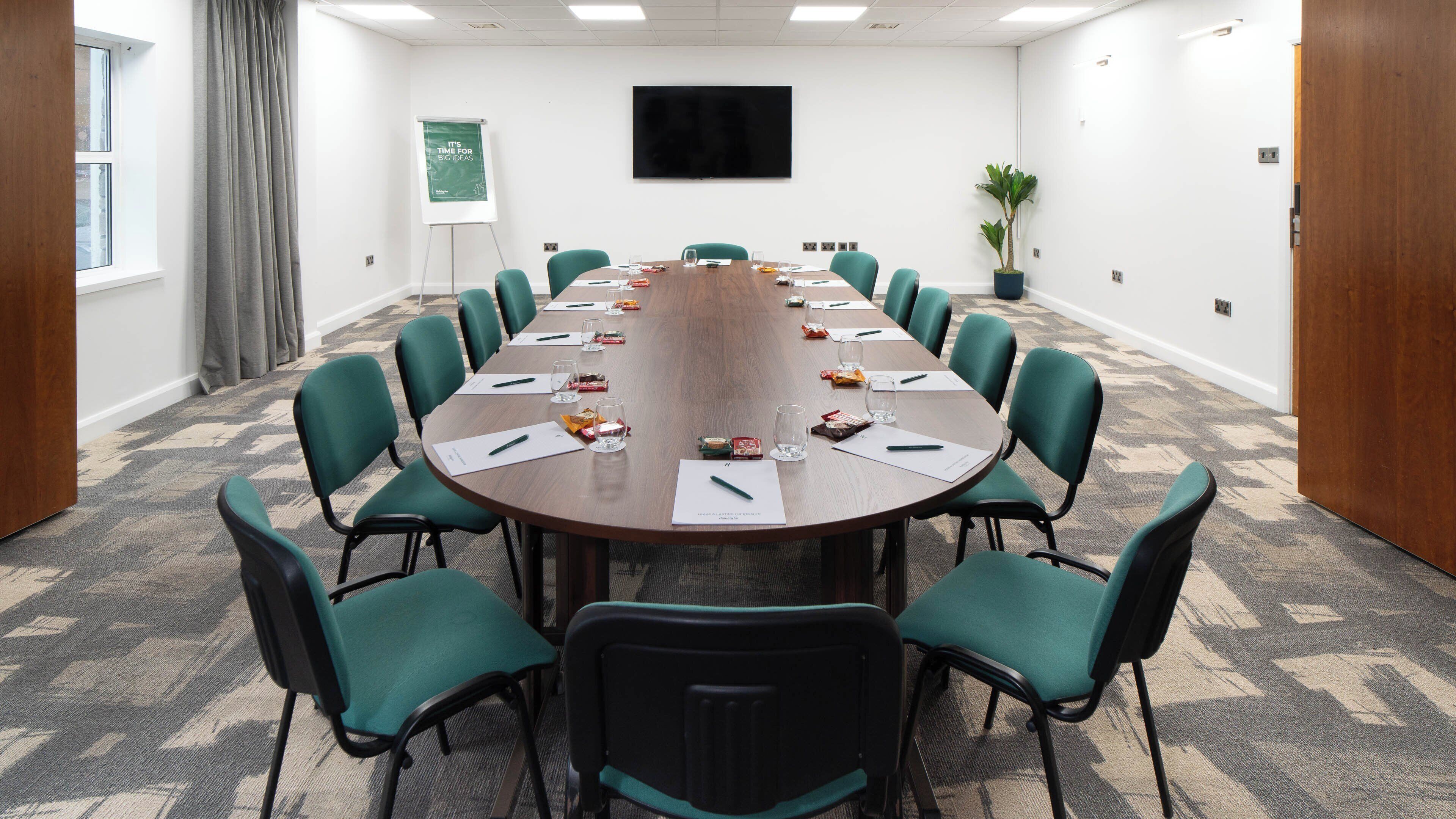 A conference room with a long table, green chairs, a whiteboard, and a TV on the wall.