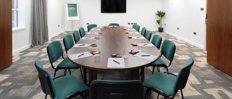 A conference room with a long table, green chairs, a whiteboard, and a TV on the wall.