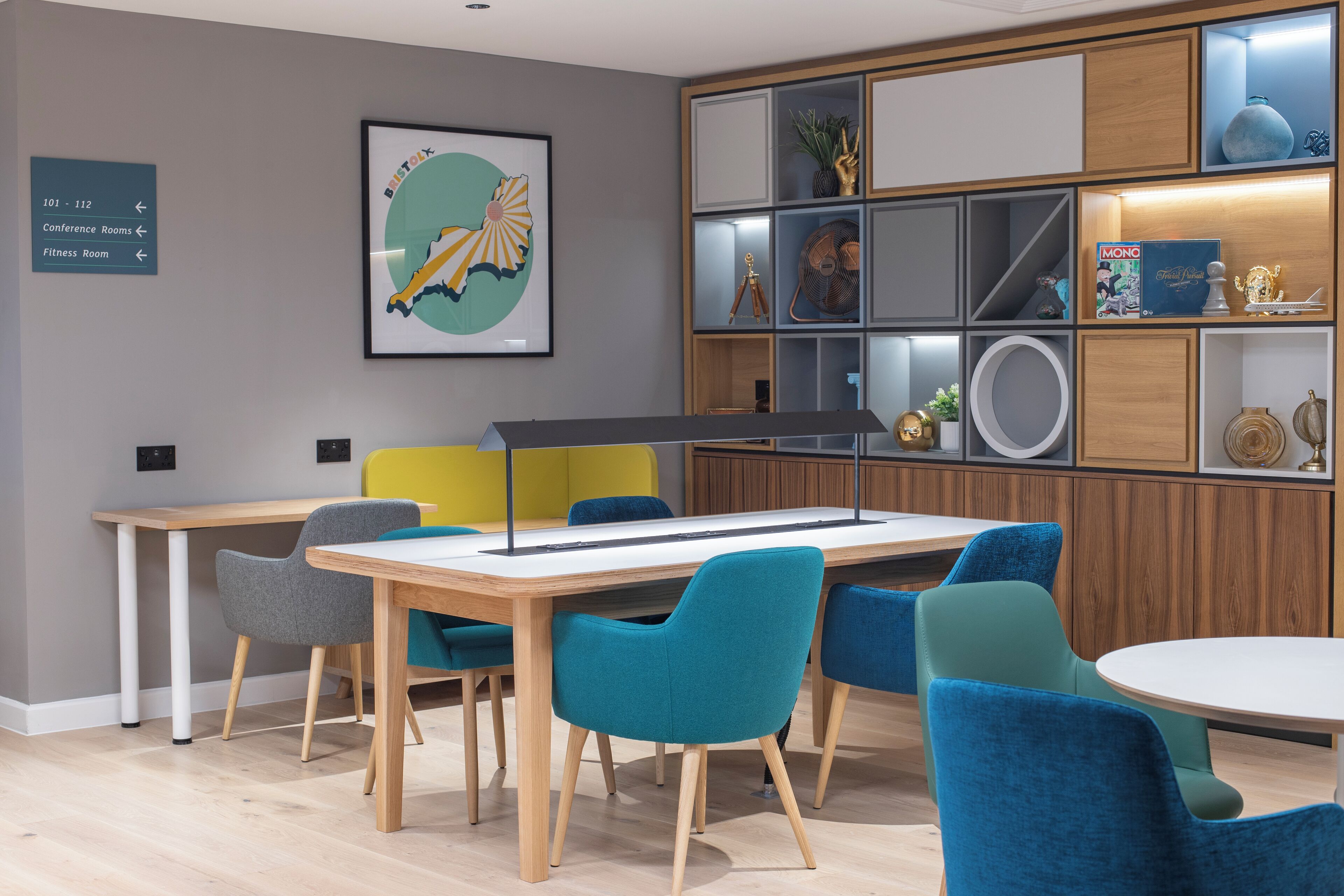 A modern conference room with a long table, teal and gray chairs, and a wooden cabinet with decorative items.