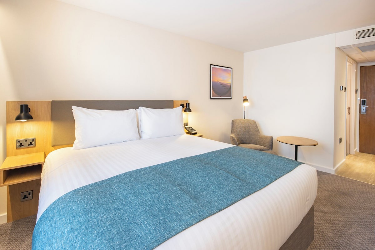 A hotel room with a large bed, bedside tables, a chair, a small table, and a door.