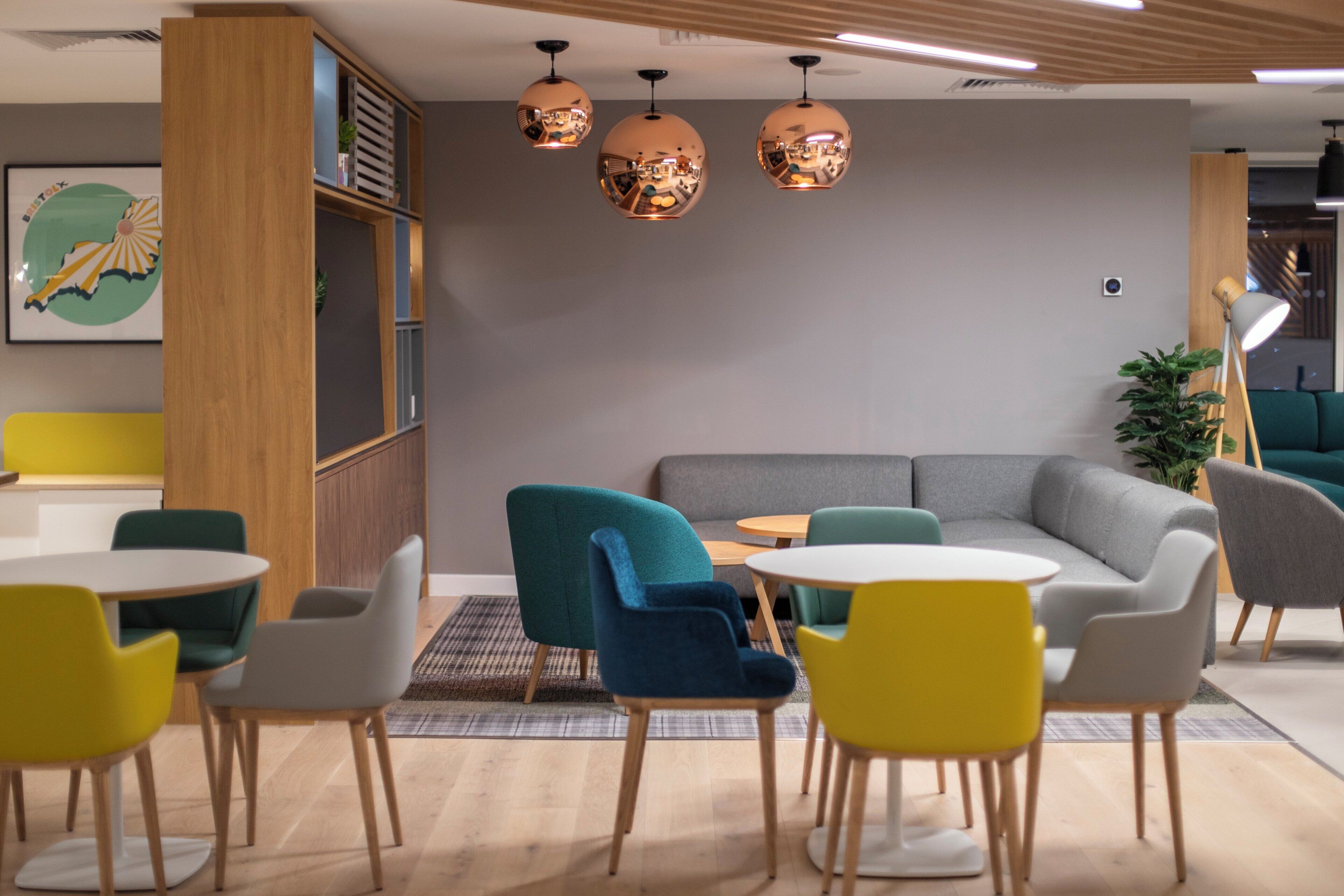 A modern lounge area with round tables and colorful chairs, a grey sofa, and a wooden bookshelf.