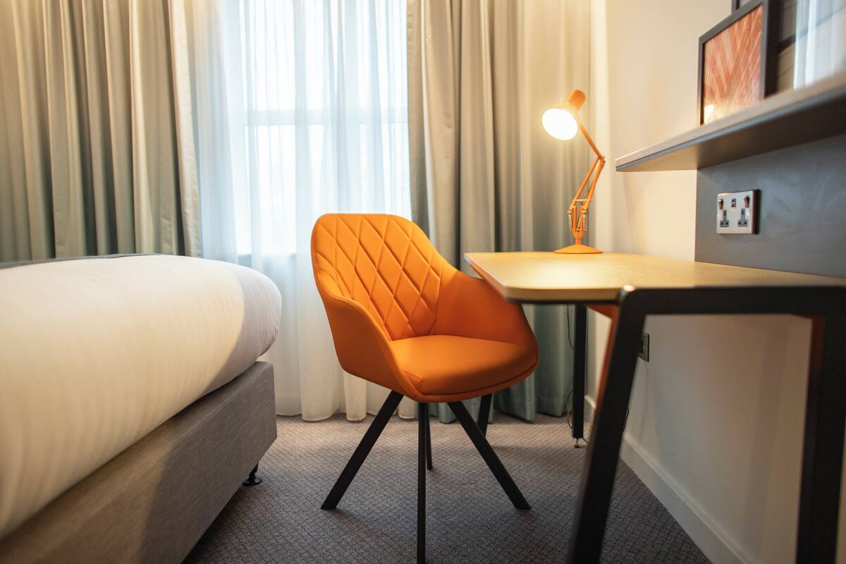A hotel room with a bed, an orange chair, a desk, a lamp, and a shelf with a picture.