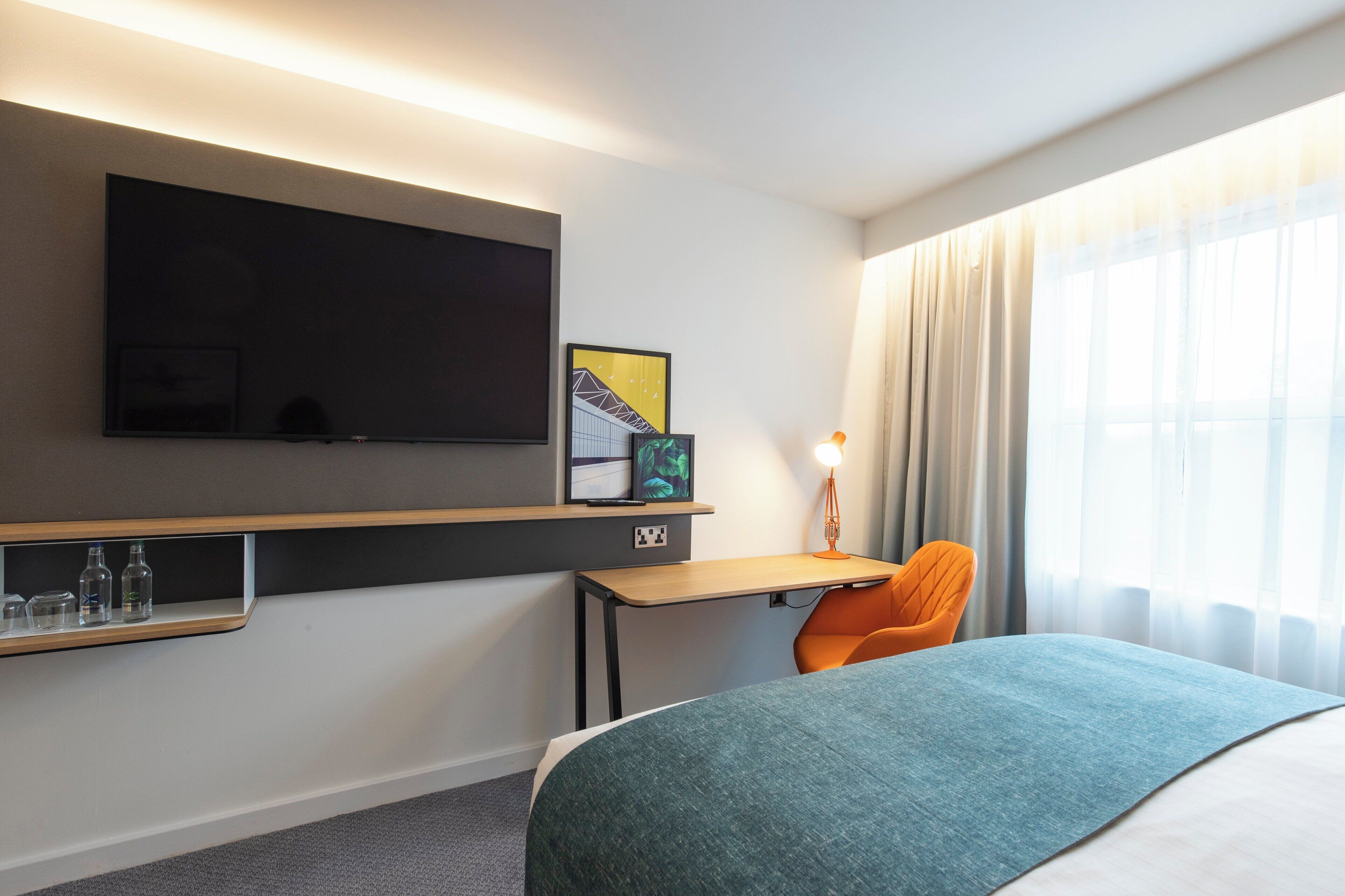 Une chambre d’hôtel comprenant un lit, un bureau, une chaise, un téléviseur fixé au mur et une fenêtre avec des rideaux.