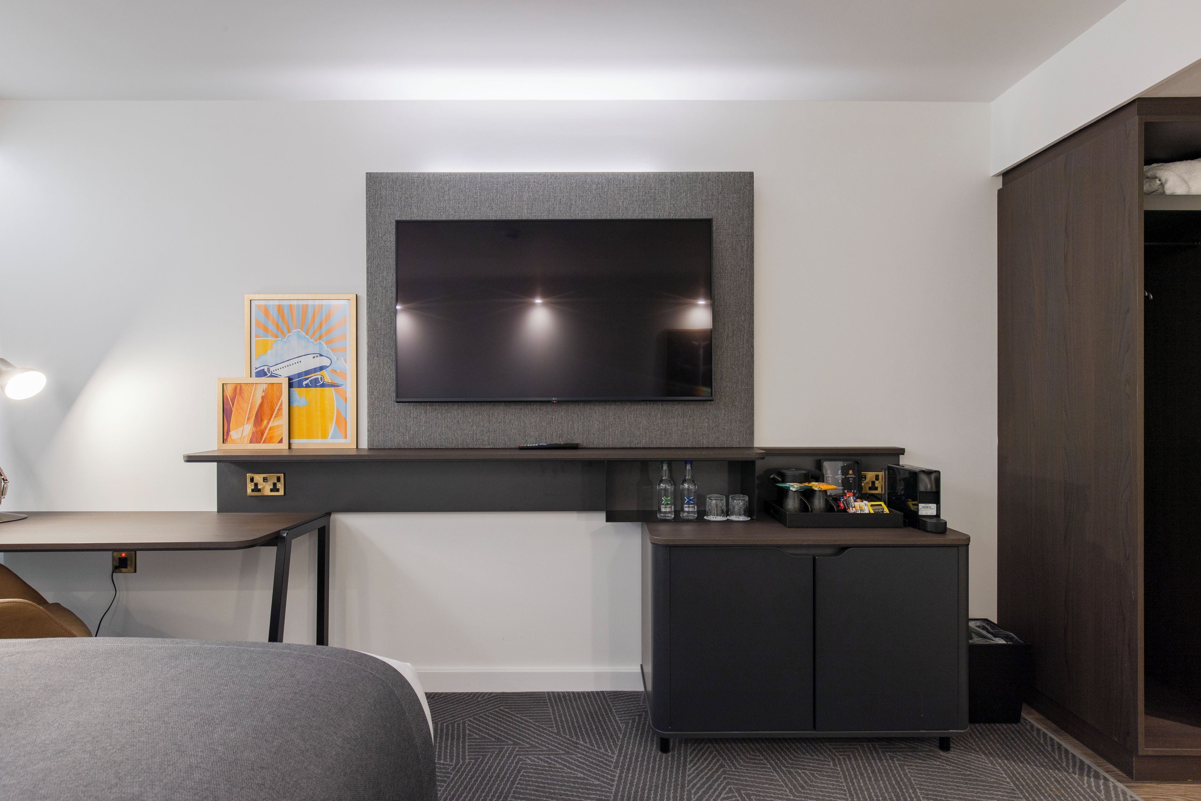 A modern hotel room with a flat-screen TV mounted on the wall, a small kitchenette with a coffee maker, and a bed with a grey headboard.