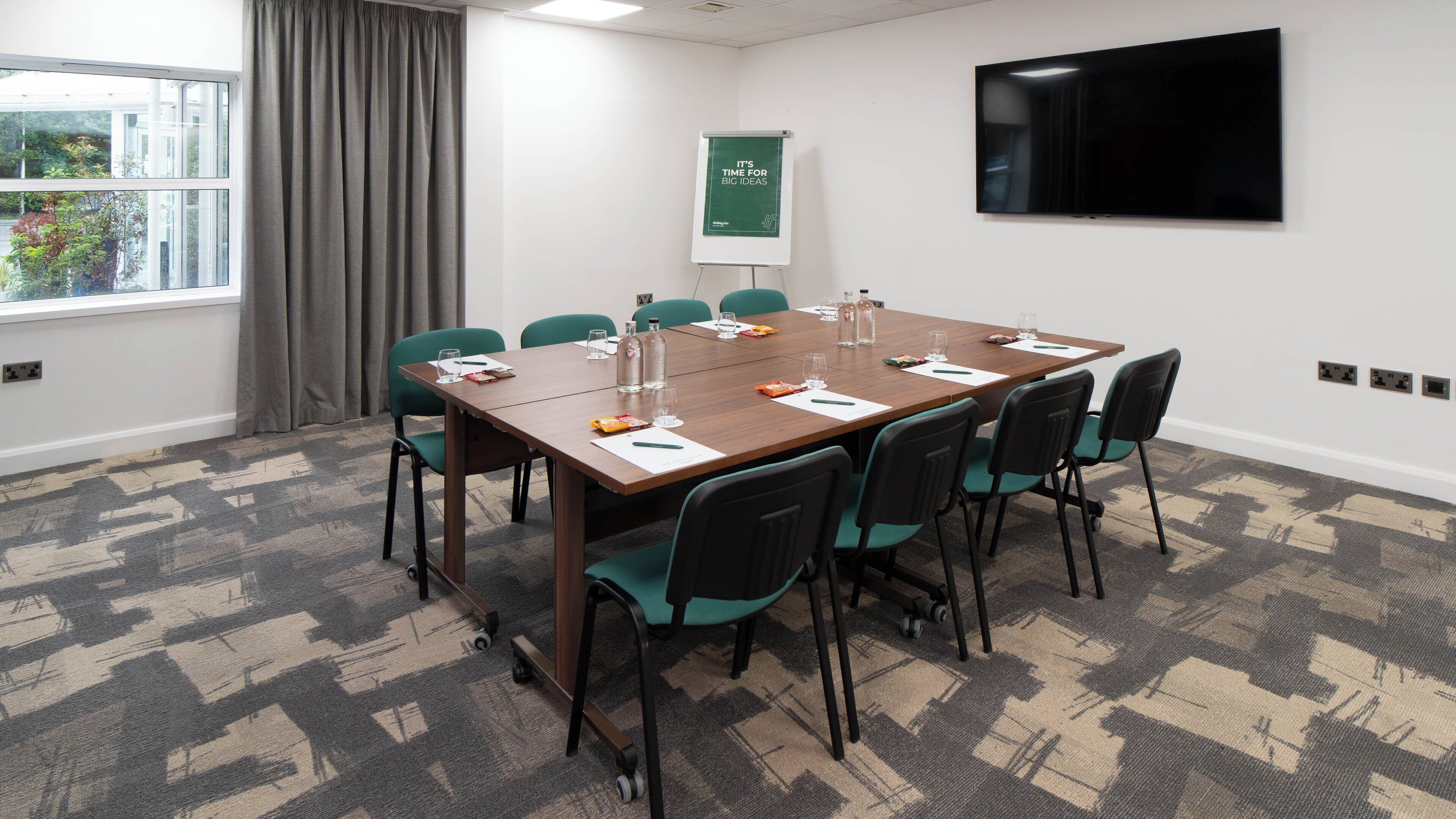 Une salle de conférence équipée d’une grande table, de chaises, d’un écran de projection et d’une fenêtre avec des rideaux.